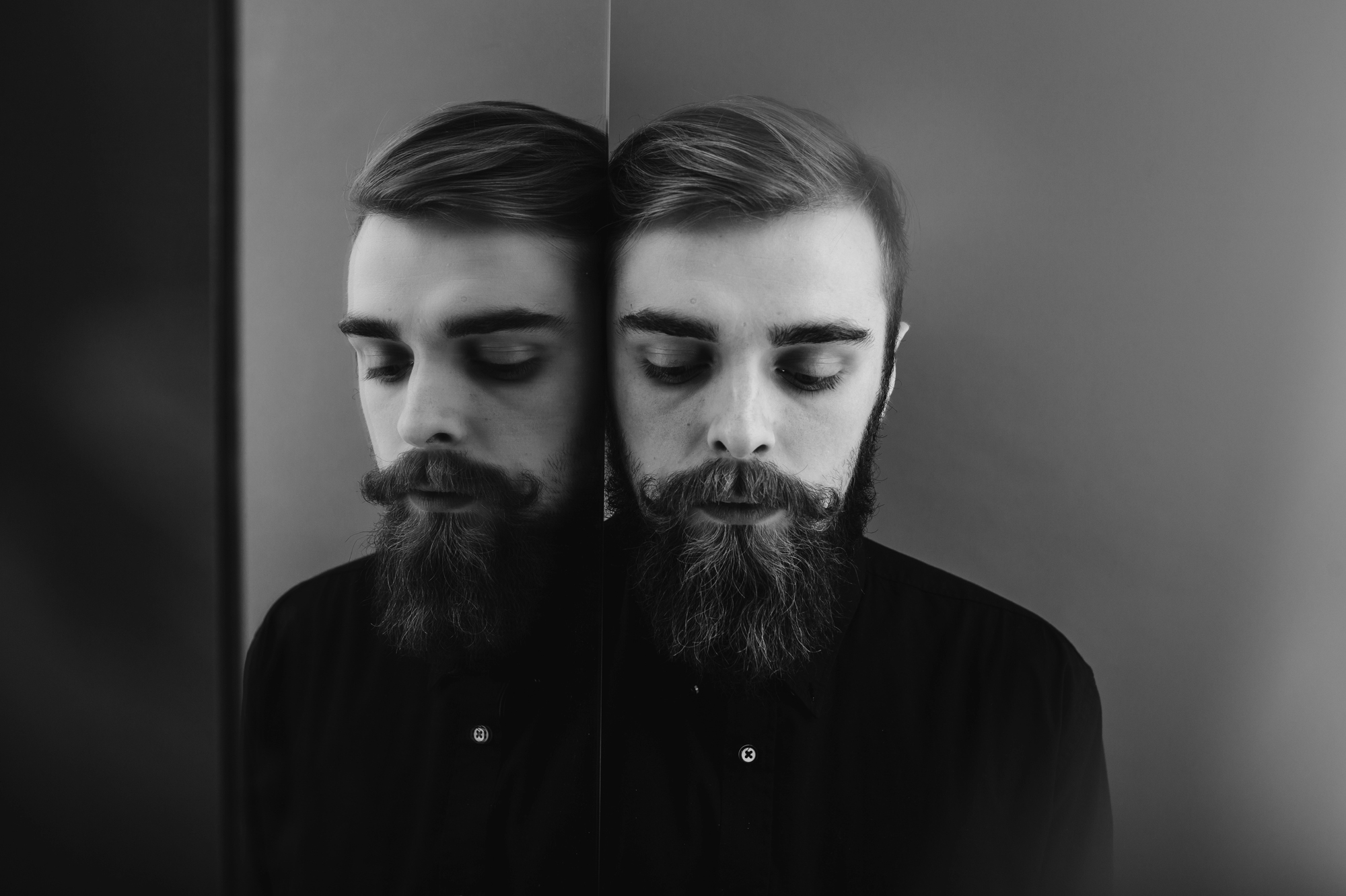 A black-and-white photo of a person with a beard and mustache, leaning against a reflective surface. The person's eyes are closed, and their reflection is clearly visible in the surface. They are wearing a dark shirt, and the background is plain.