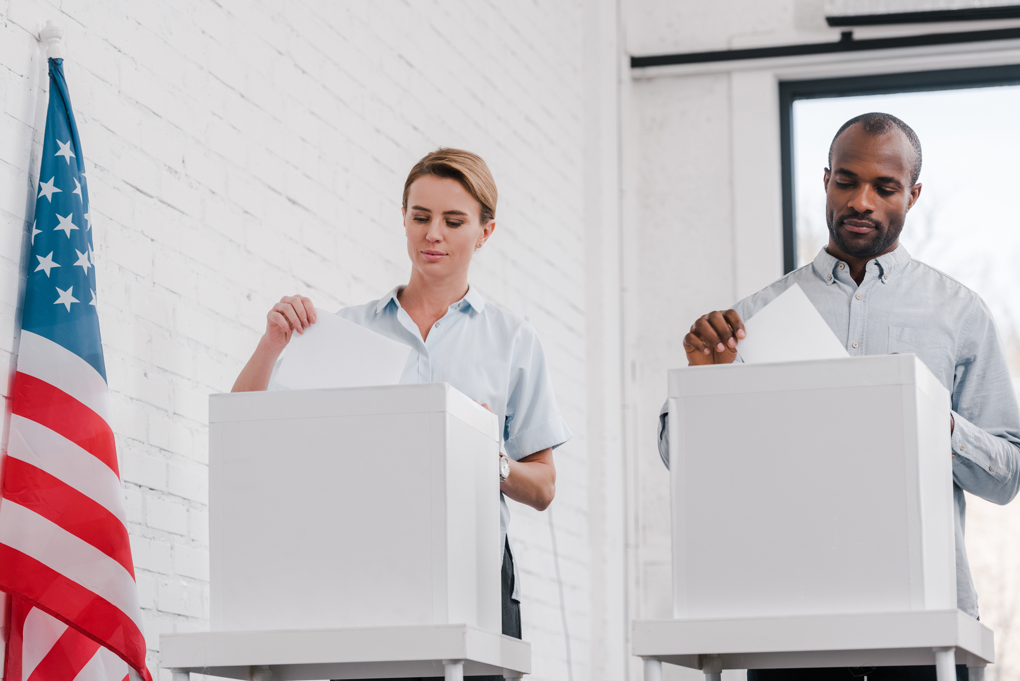 Two individuals, a woman and a man, are casting ballots at white polling booths. They are located in front of a white brick wall, with an American flag to the side. The scene appears to take place indoors, possibly in a voting center.