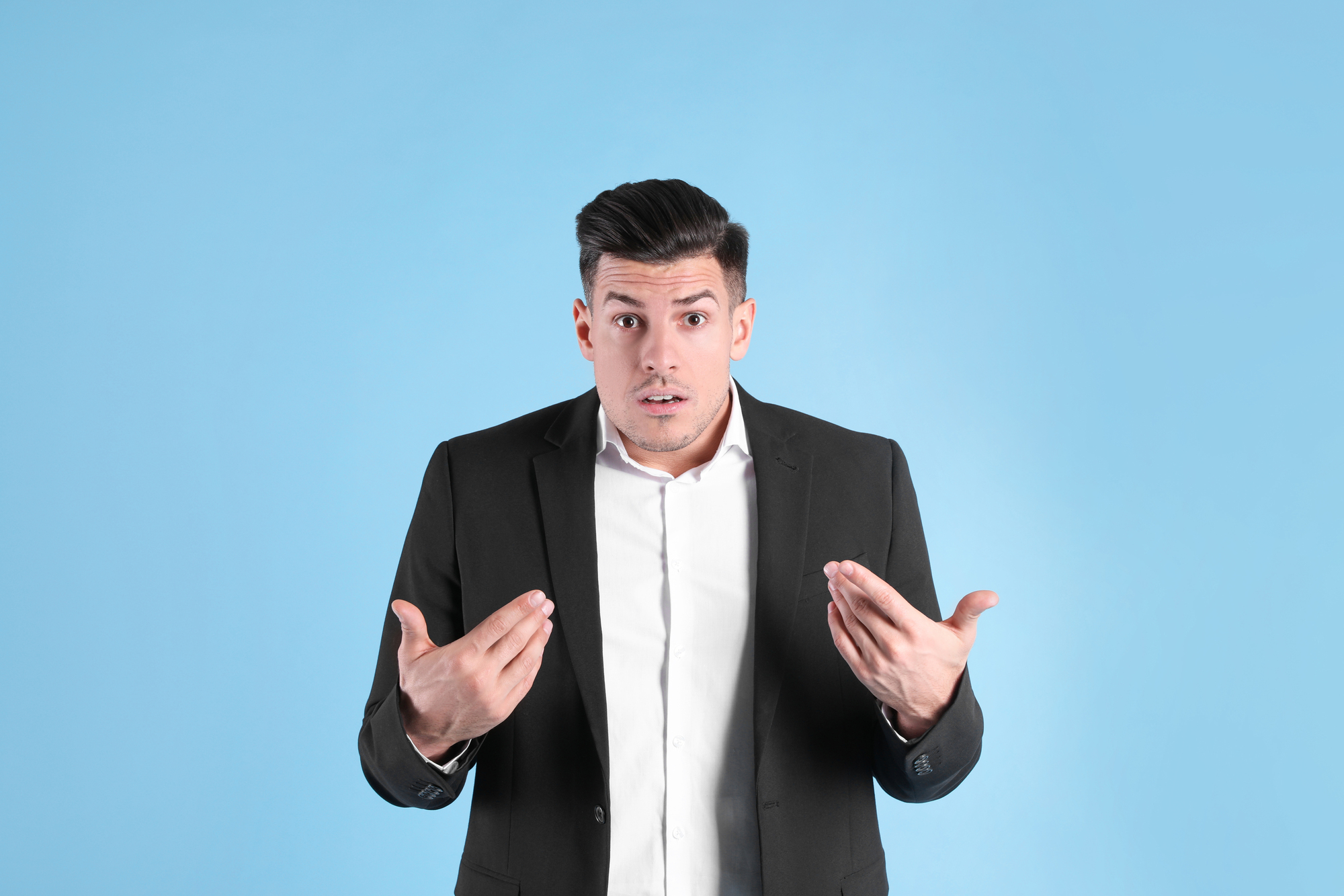 A man in a black suit and white shirt stands against a light blue background. He has a surprised expression on his face, with both hands raised and palms facing upwards.