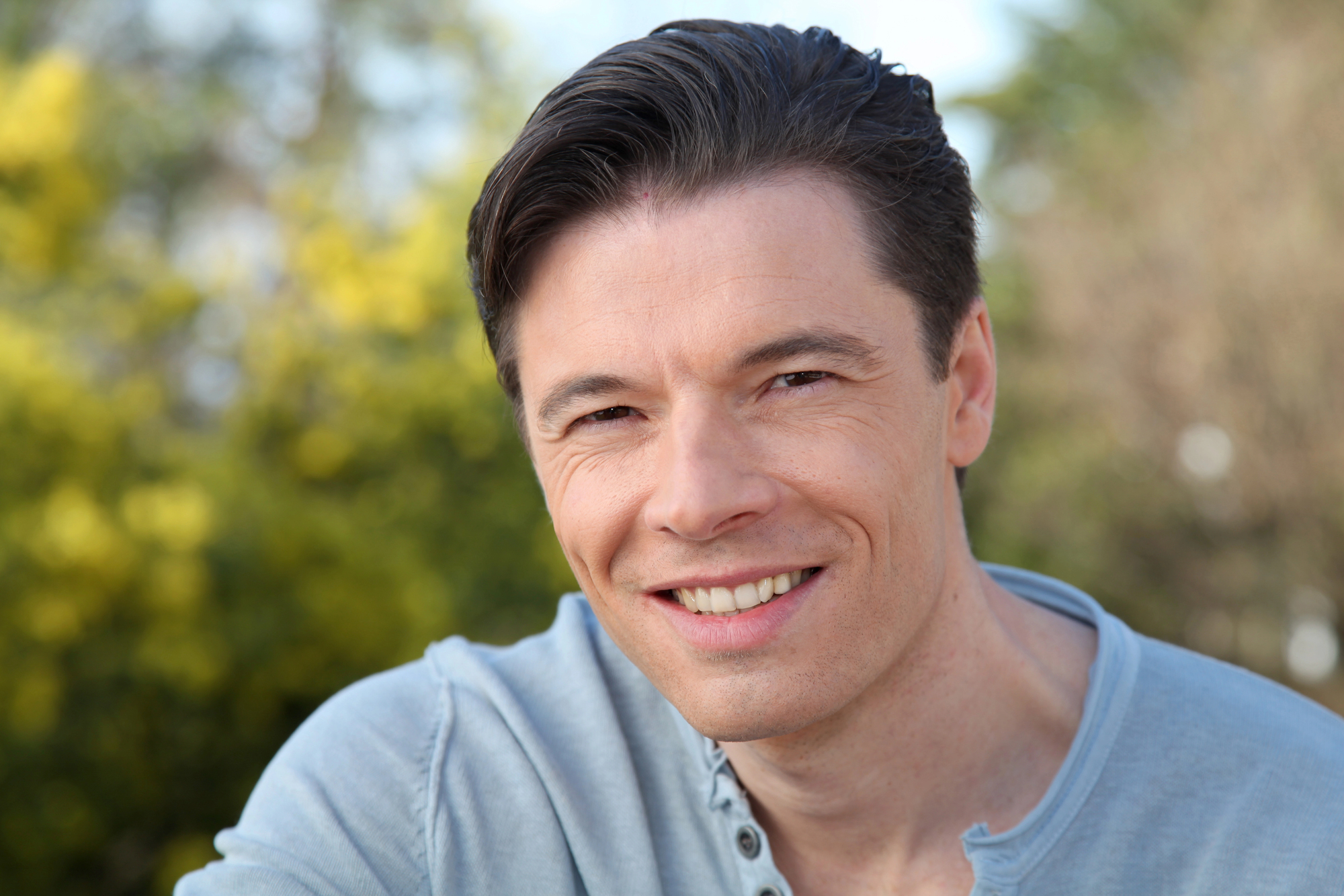 A smiling man with short dark hair is wearing a light gray shirt with buttons near the collar. He is standing outdoors with blurred greenery and yellow flowers in the background.