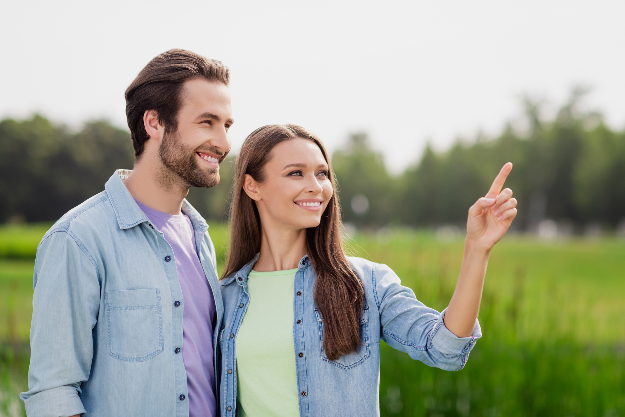 A smiling man and woman stand side by side outdoors, with lush green trees and grass in the background. Both are wearing casual denim shirts. The woman points at something in the distance while the man looks in the same direction.