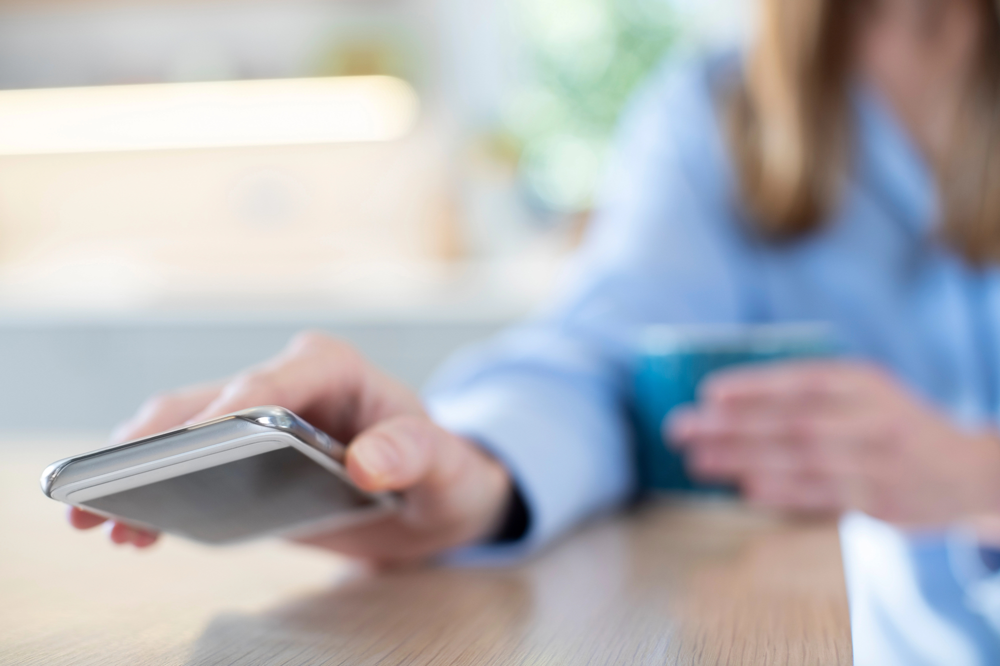 A person in a blue shirt holds a smartphone in one hand and a teal coffee mug in the other. The background is blurred and richly lit, suggesting a kitchen or living space. The focus is on the smartphone and the person's hand.