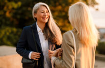 Two women are engaging in a friendly conversation outdoors. One woman with long gray hair is smiling brightly, dressed in a navy blazer and white shirt, carrying a black shoulder bag. The other woman is seen from behind, with blonde hair, wearing a tan blazer.