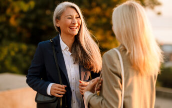 Two women are engaging in a friendly conversation outdoors. One woman with long gray hair is smiling brightly, dressed in a navy blazer and white shirt, carrying a black shoulder bag. The other woman is seen from behind, with blonde hair, wearing a tan blazer.