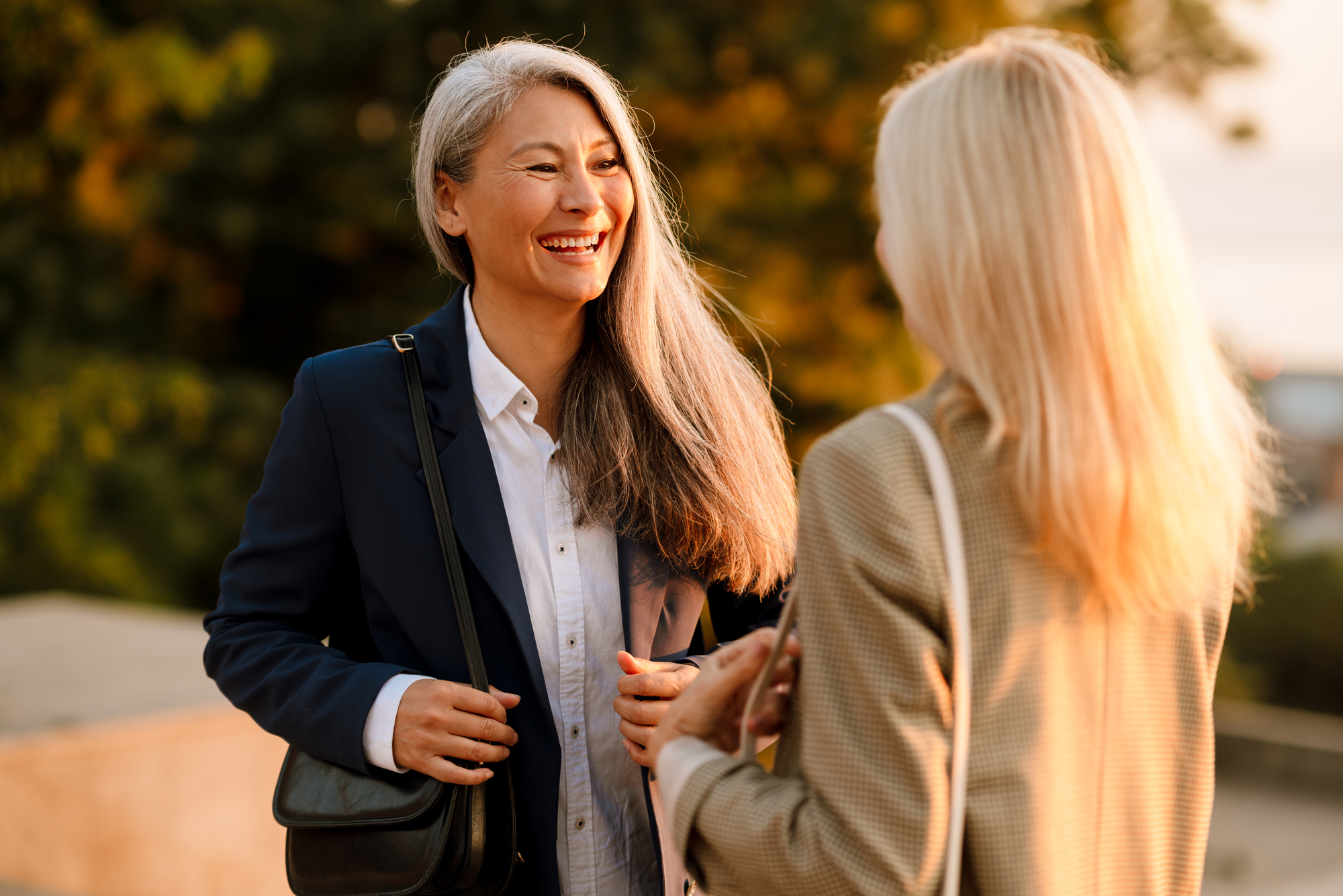 Two women are engaging in a friendly conversation outdoors. One woman with long gray hair is smiling brightly, dressed in a navy blazer and white shirt, carrying a black shoulder bag. The other woman is seen from behind, with blonde hair, wearing a tan blazer.