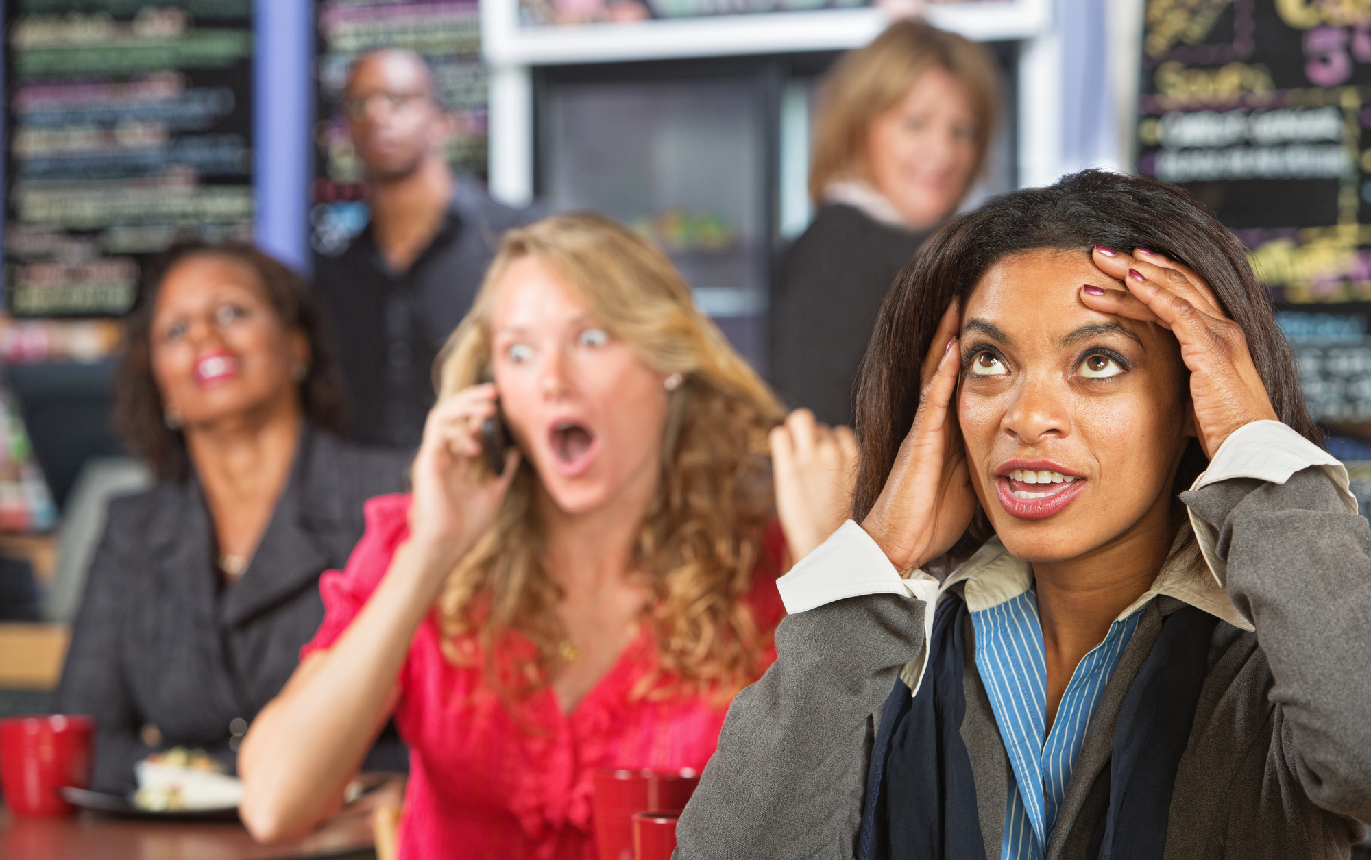 A woman in the foreground has her hands on her head, looking concerned. Behind her, a blonde woman is on her phone with a shocked expression. Another woman in red is smiling, and a man and woman are in the background, slightly blurred.