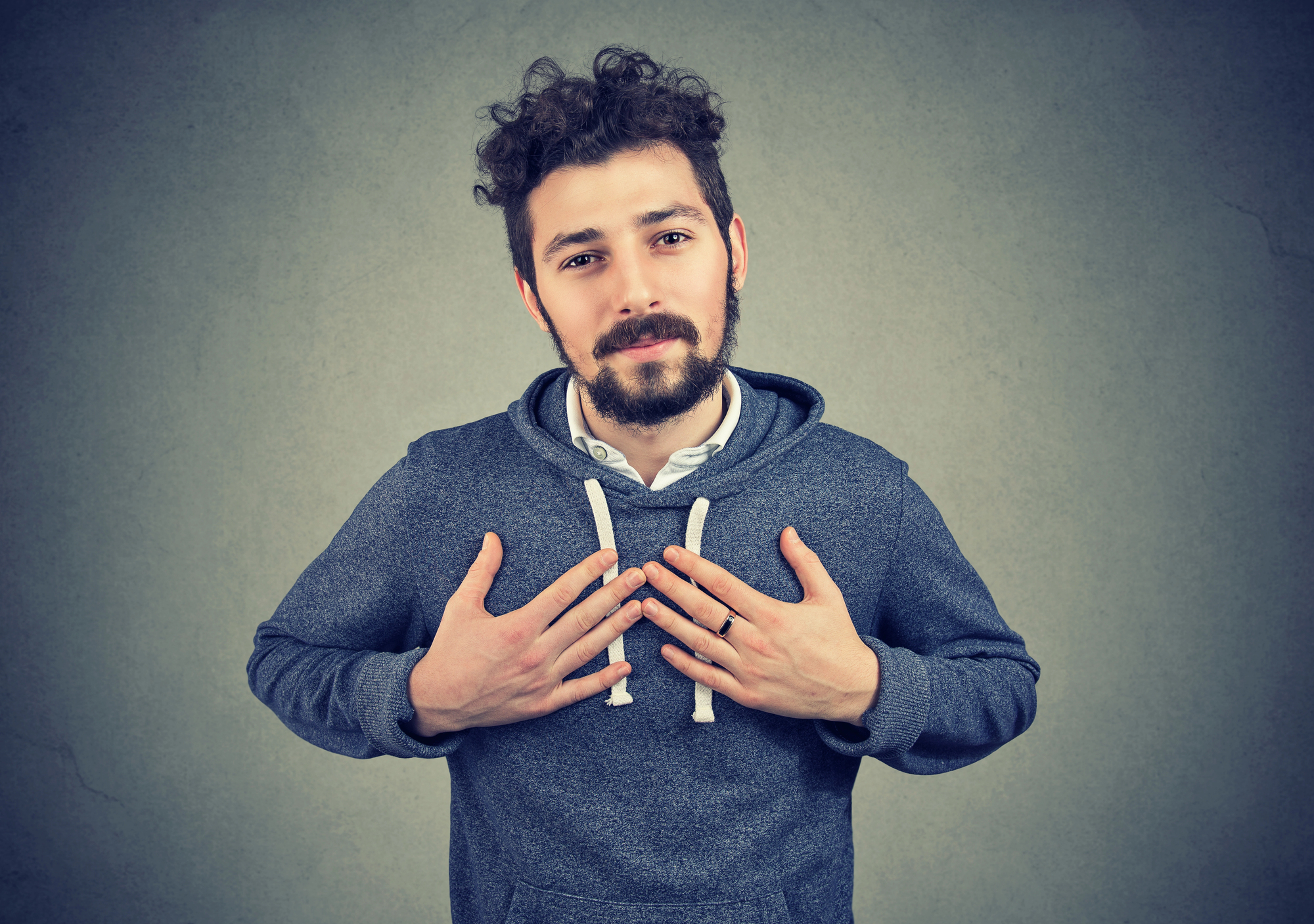 A man with curly hair and a beard stands against a gray background. He is wearing a gray hoodie and has his hands placed on his chest, projecting a thoughtful or apologetic expression. His gaze is directed towards the camera.