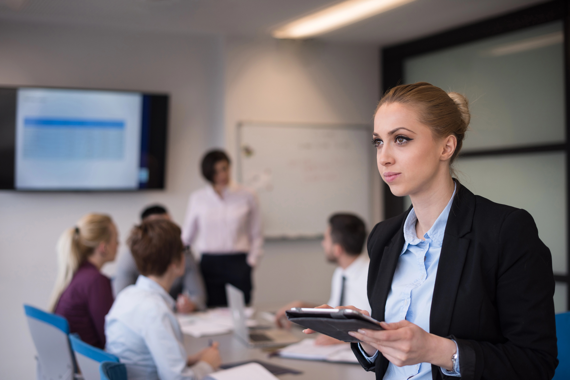 A woman stands in the foreground, holding a tablet, and appears focused. In the background, four people sit around a table with laptops, and another person stands near a whiteboard, giving a presentation. A wall-mounted screen displays a slide in the meeting room.