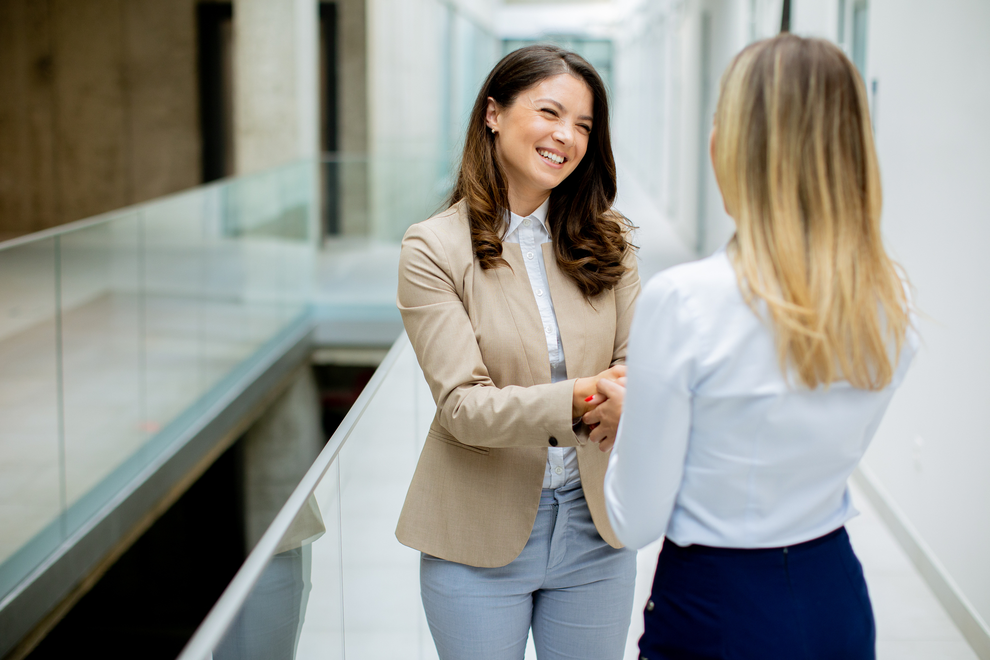 Two women standing and shaking hands in a bright, modern hallway. One woman, with long dark hair and dressed in a beige blazer, is smiling. The other woman, with long blonde hair, is seen from the back, wearing a white shirt and dark skirt.