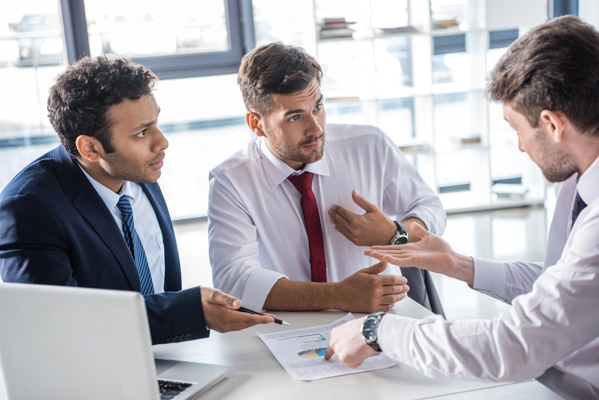 Three men in business attire are engaged in a serious discussion around a table. One points to a document with charts, while the others listen attentively. A laptop is open on the table, and a bright office with shelves in the background.