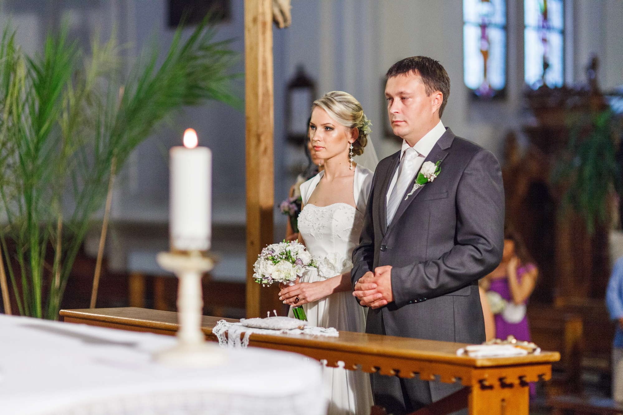 A bride and groom stand solemnly at the altar during a wedding ceremony. The bride holds a bouquet of white flowers and wears a white dress, while the groom is in a gray suit with a white boutonniere. An altar candle burns in the foreground, and greenery decorates the background.