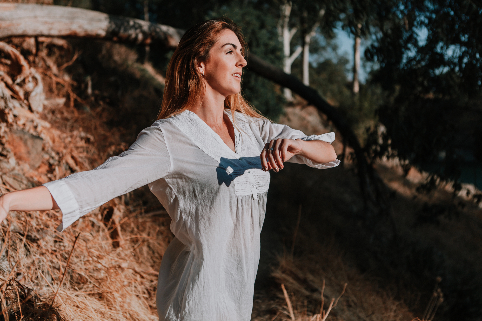 A woman with long, light brown hair stands outdoors with her arms gracefully extended. She is wearing a loose white dress and appears to be enjoying the natural surroundings, which include dry grass and trees. The sunlight casts a warm glow on her and the scenery.