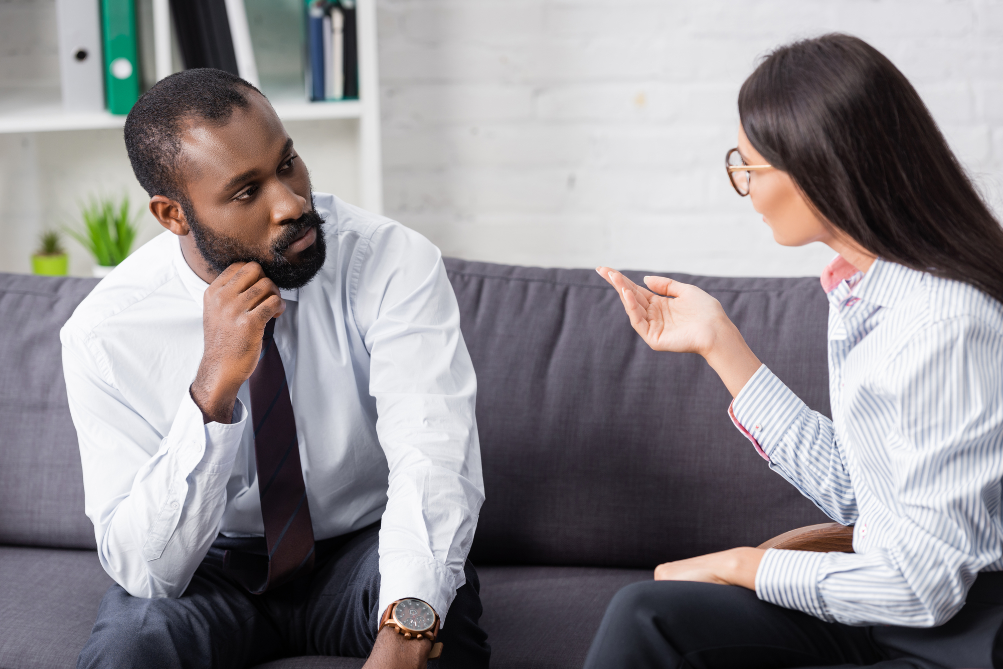 A man and a woman are sitting and having a serious conversation on a gray sofa. The man, wearing a white shirt and tie, is listening intently with his hand on his chin. The woman, wearing glasses and a striped shirt, is gesturing with her hand while speaking.