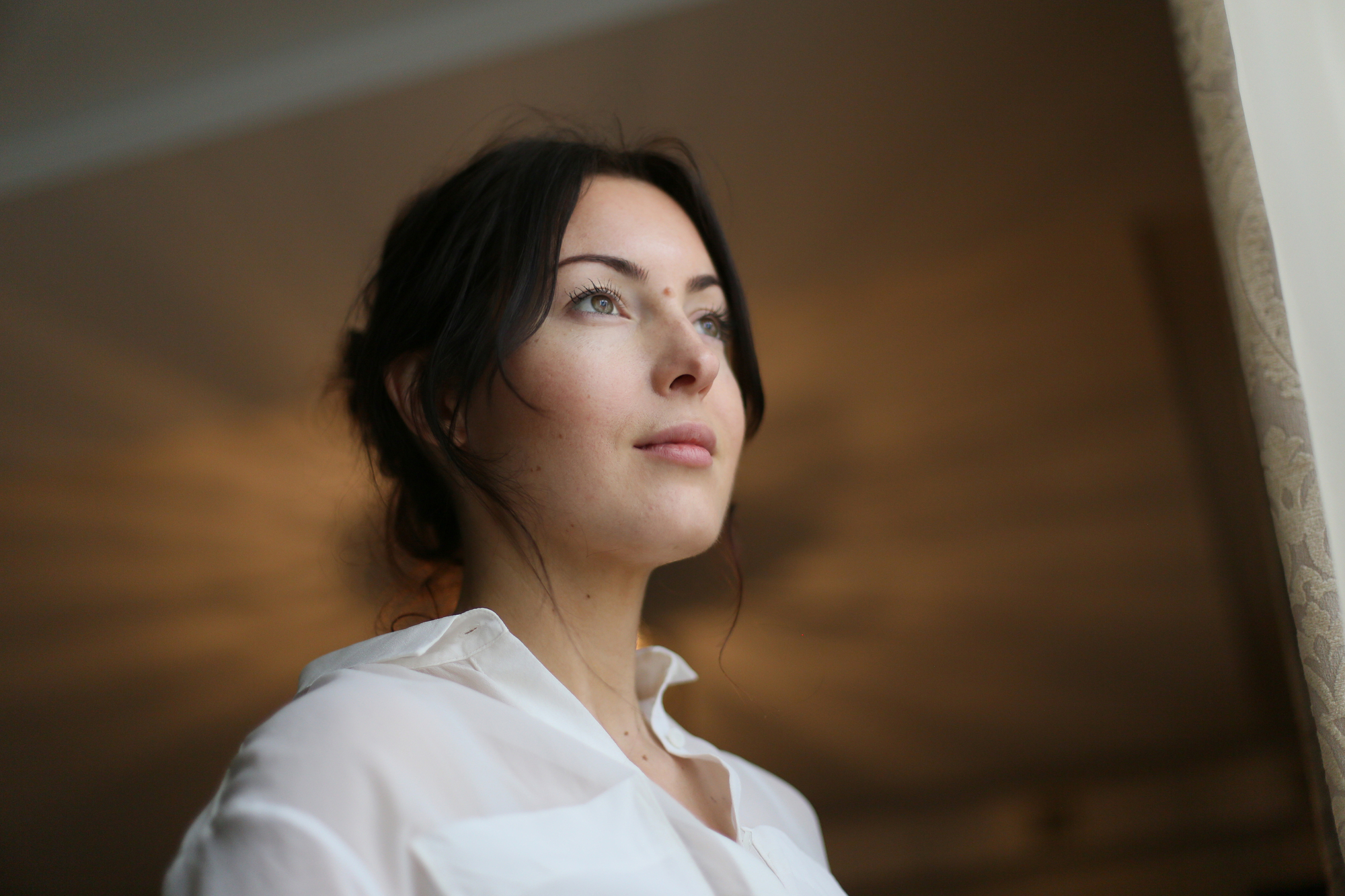 A woman with dark hair tied back stands in front of a softly lit background, looking slightly upwards and to the side. She is wearing a white shirt and has a thoughtful expression on her face. The lighting creates a warm, gentle glow around her.