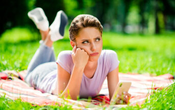 A woman lies on a red and white checkered picnic blanket on a grassy lawn, propping her head up with her left hand while holding a smartphone in her right hand. She gazes to the left with a pensive or slightly annoyed expression, resting on her stomach.