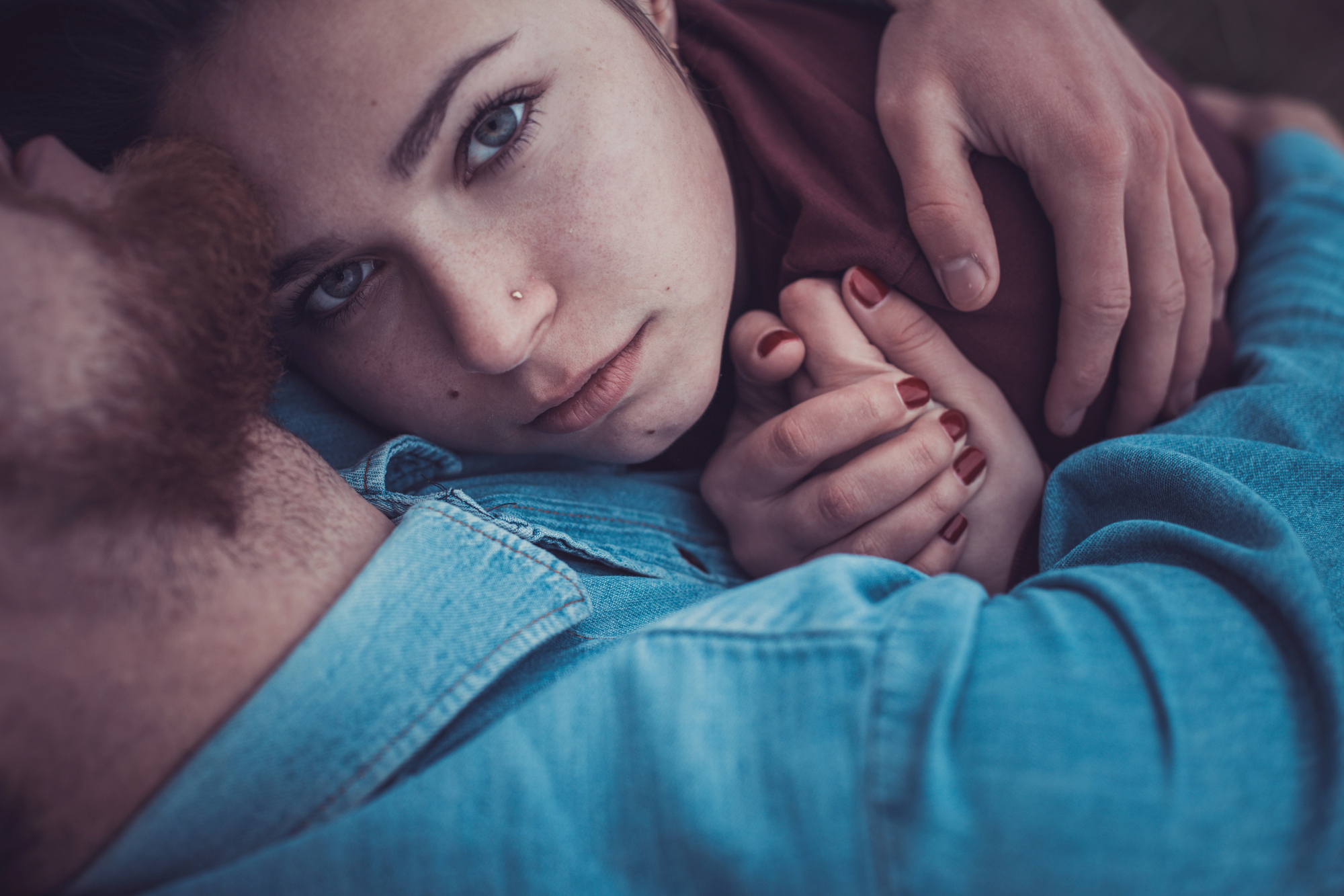 A close-up of two people embracing. One person, with piercing eyes and a nose ring, looks directly at the camera while lying against the other, who has a beard. Hands are intertwined, with maroon-painted nails visible. Both wear denim-like fabric.