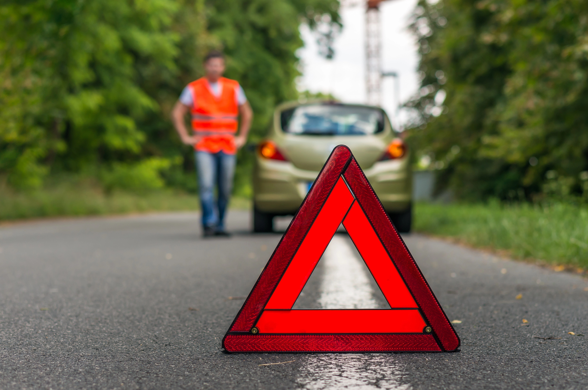 A red warning triangle is placed on the road, indicating a breakdown. In the background, a person wearing a high-visibility vest stands near a parked car with the trunk open. The scene is set on a tree-lined road.