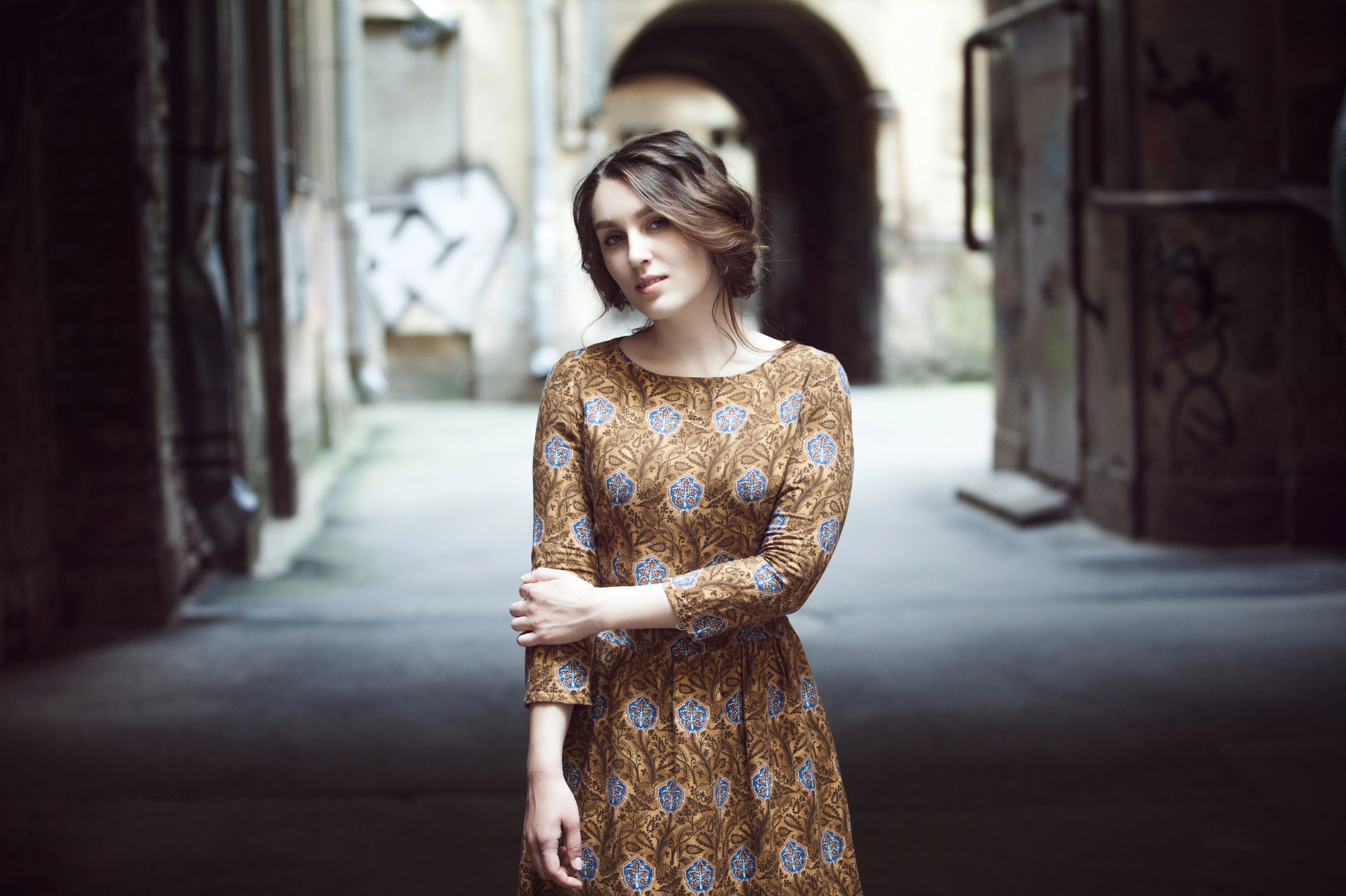 A woman with wavy hair, wearing a long-sleeved, patterned dress, stands in a slightly shadowed, urban alleyway with graffiti on the walls. She gazes at the camera with one arm crossed and the other hand resting on her arm.