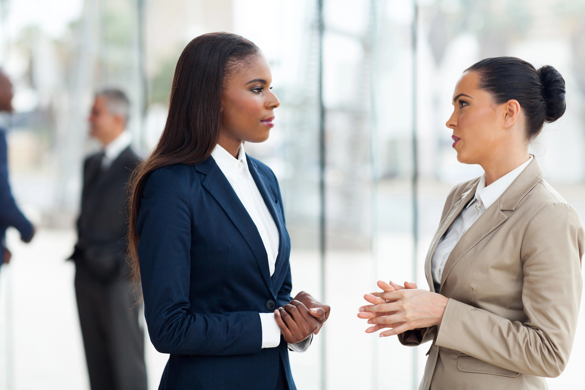 Two businesswomen in professional attire, one in a navy suit and the other in a beige suit, are engaged in a conversation in a bright, modern office space. In the blurred background, two men are also conversing near a window with a city view.