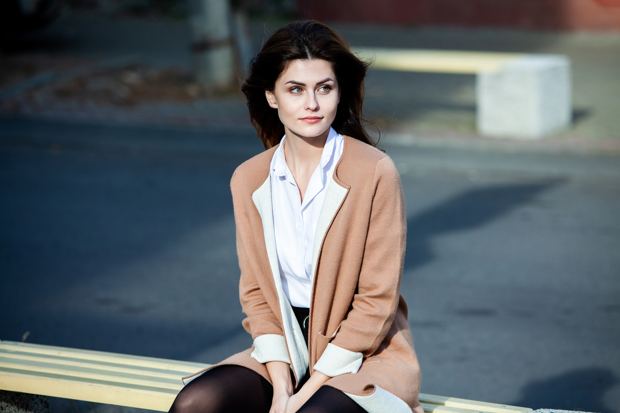 A young woman with long brown hair and dressed in a white blouse and beige coat sits on a yellow bench outdoors. She gazes off to the side with a contemplative expression. The background shows an urban street setting.