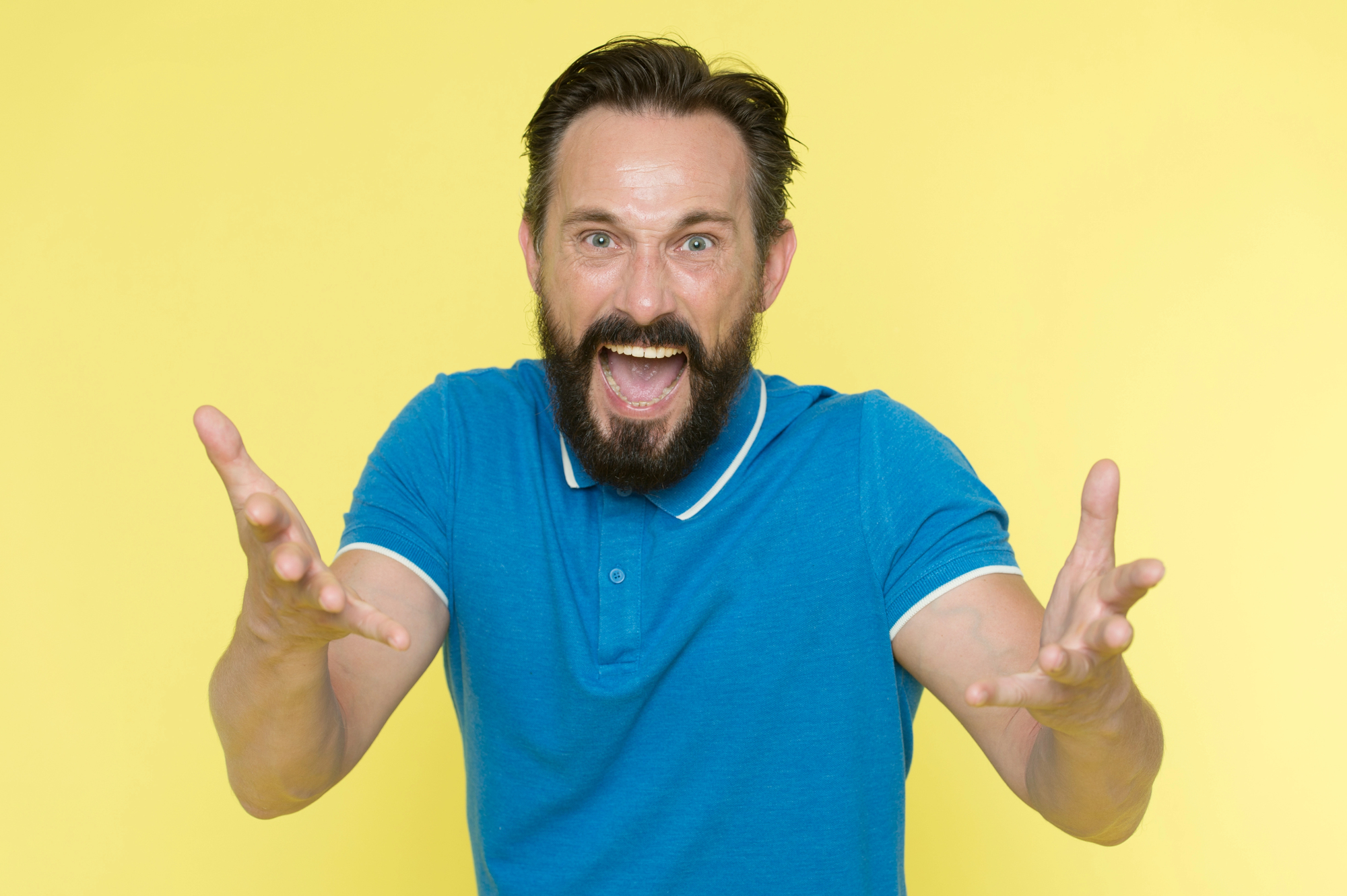 A man with a beard and mustache, wearing a blue polo shirt, is smiling broadly with his mouth open, extending both arms towards the camera. The background is a solid yellow color.