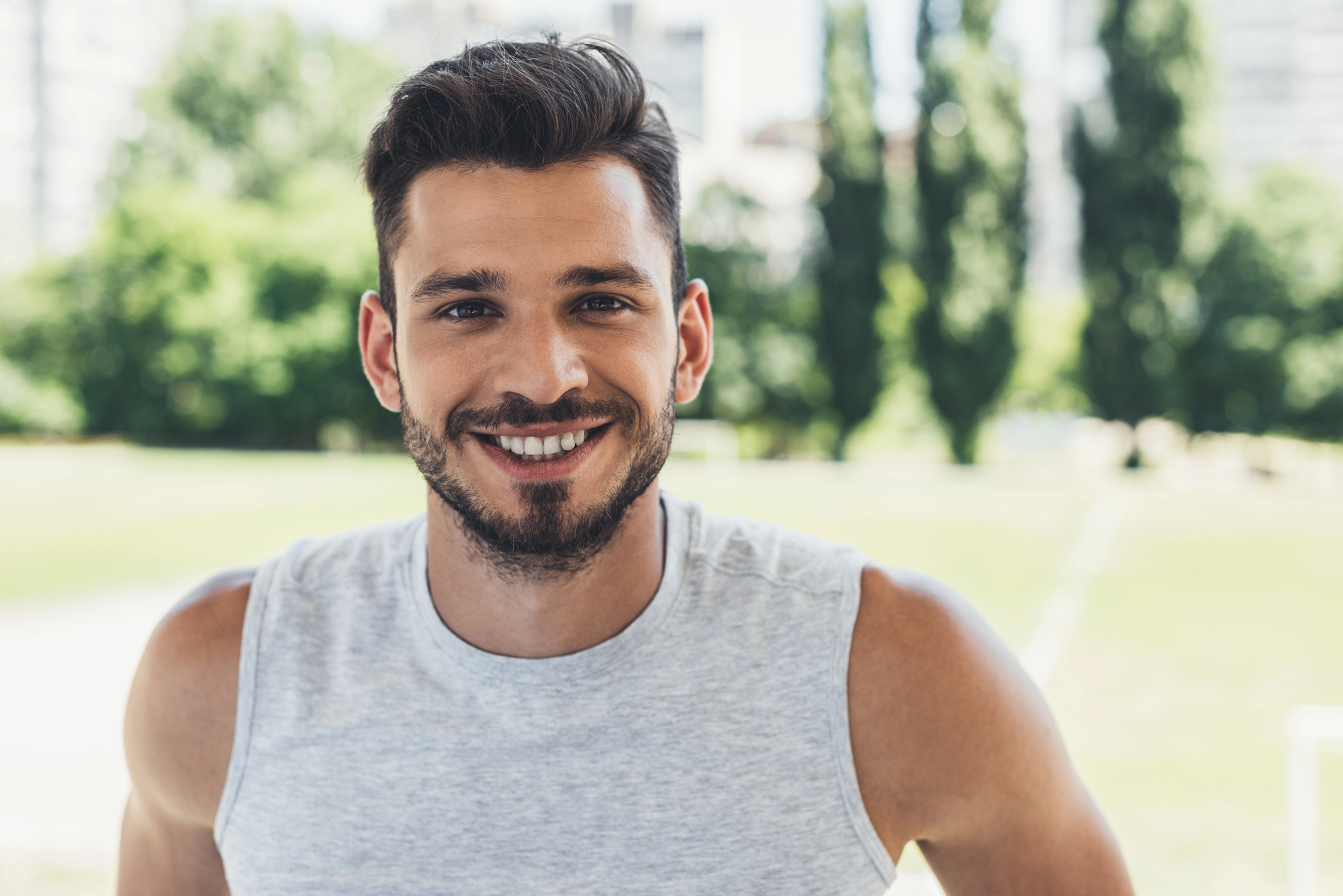 A smiling man with short, dark hair and a beard is wearing a sleeveless gray shirt. He is standing outdoors in a park with green trees and blurred buildings in the background, under bright daylight.