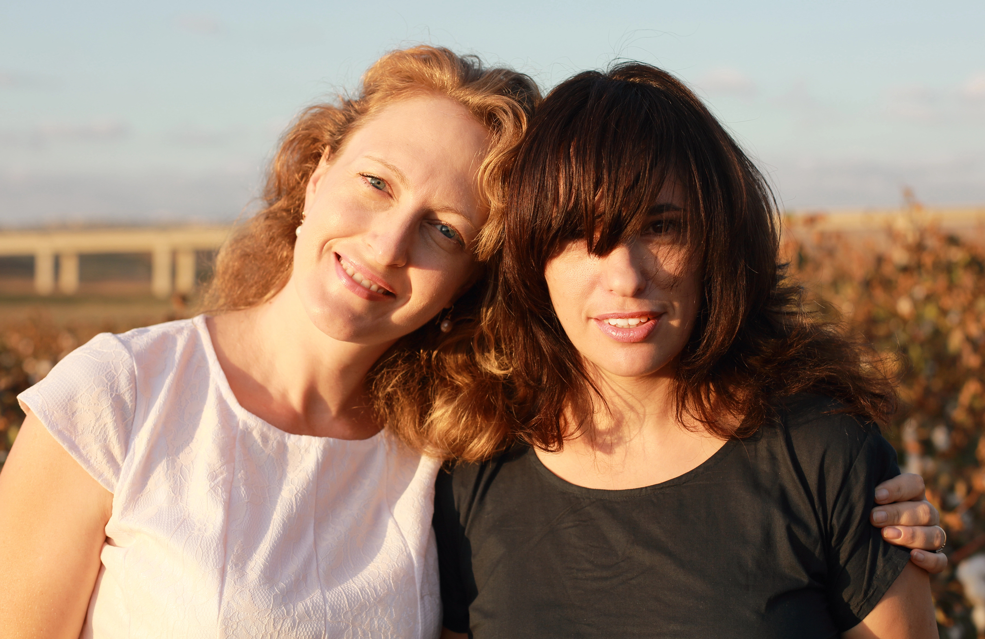 Two women stand outdoors, smiling at the camera. The woman on the left has light curly hair and is wearing a white top, while the woman on the right has dark hair with bangs and is wearing a black top. They have their arms around each other, with a scenic background behind them.