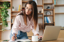 A woman with long brown hair and glasses is leaning over a desk, writing in a notebook with a pen while looking at an open laptop. She is dressed in a light pink shirt and jeans. The desk has a white coffee mug and a smartphone. Shelves with books and office items are in the background.