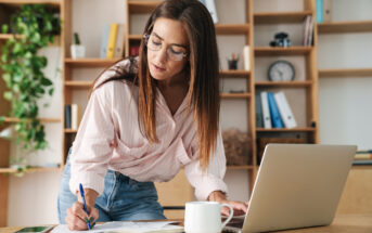 A woman with long brown hair and glasses is leaning over a desk, writing in a notebook with a pen while looking at an open laptop. She is dressed in a light pink shirt and jeans. The desk has a white coffee mug and a smartphone. Shelves with books and office items are in the background.