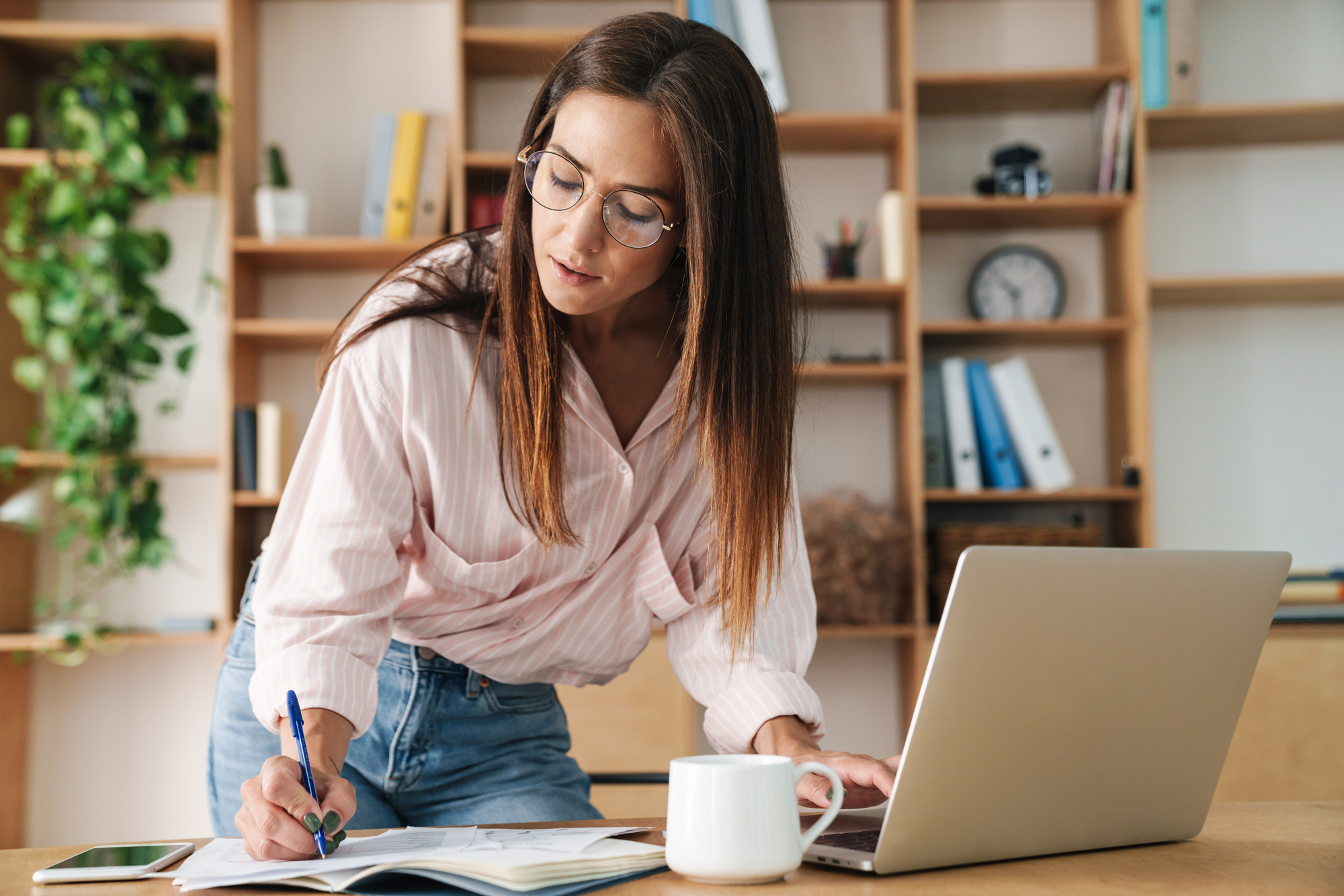 A woman with long brown hair and glasses is leaning over a desk, writing in a notebook with a pen while looking at an open laptop. She is dressed in a light pink shirt and jeans. The desk has a white coffee mug and a smartphone. Shelves with books and office items are in the background.