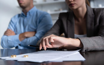 A man and a woman sit at a table, both looking serious. The man has his arms crossed while the woman, holding a pen, gestures towards a document in front of them. Two golden rings lie on the table, indicating a possible discussion about divorce or separation.