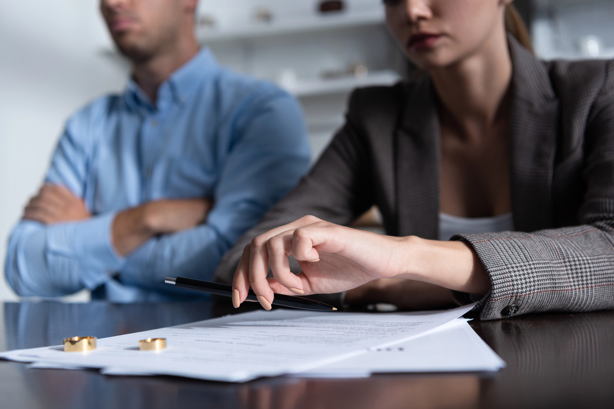 A man and a woman sit at a table, both looking serious. The man has his arms crossed while the woman, holding a pen, gestures towards a document in front of them. Two golden rings lie on the table, indicating a possible discussion about divorce or separation.