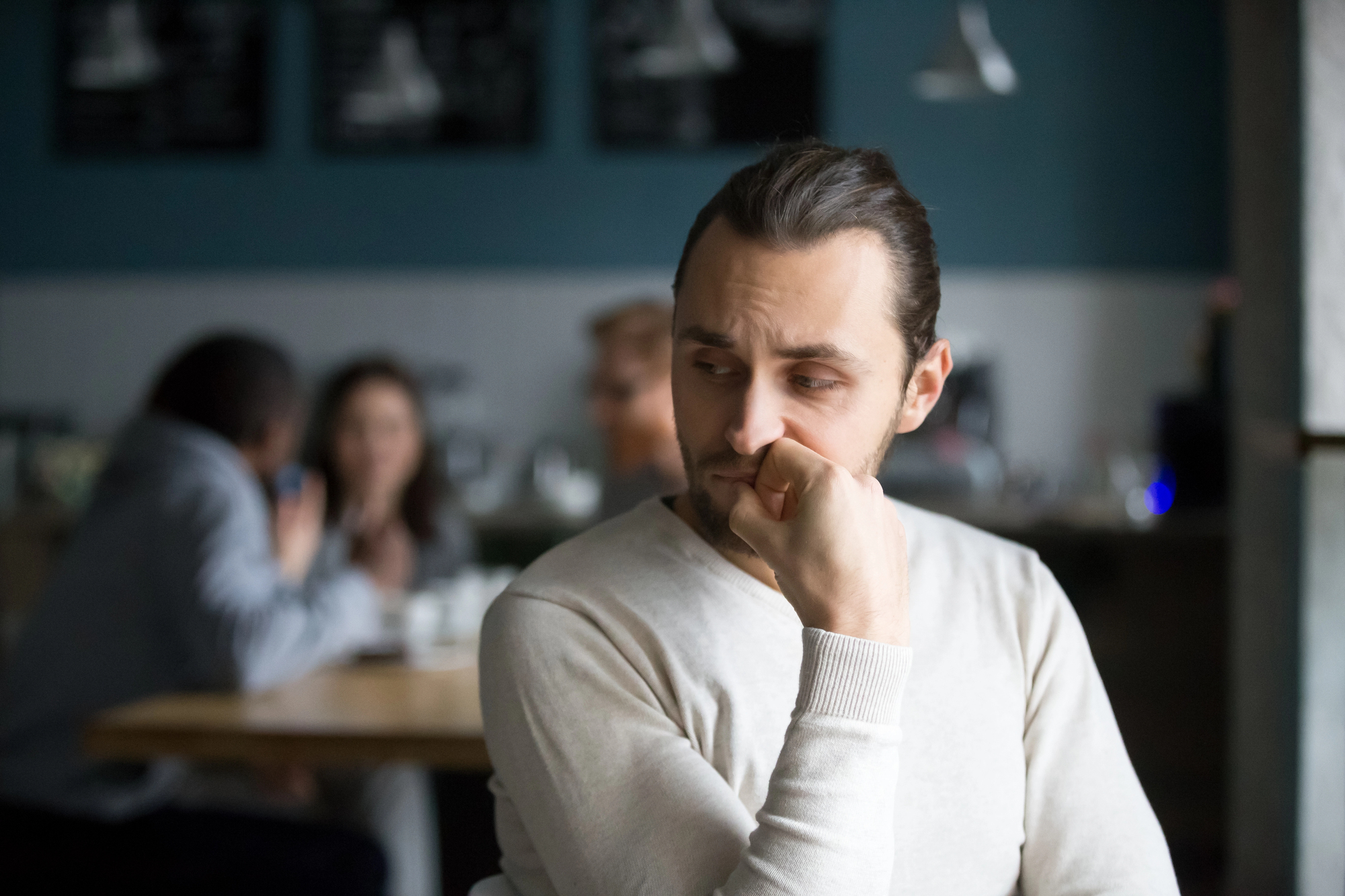 A man with a serious expression is sitting alone at a café table, resting his chin on his hand. In the background, a group of people are conversing and laughing. The café has a casual atmosphere with dim lighting and blurred details.