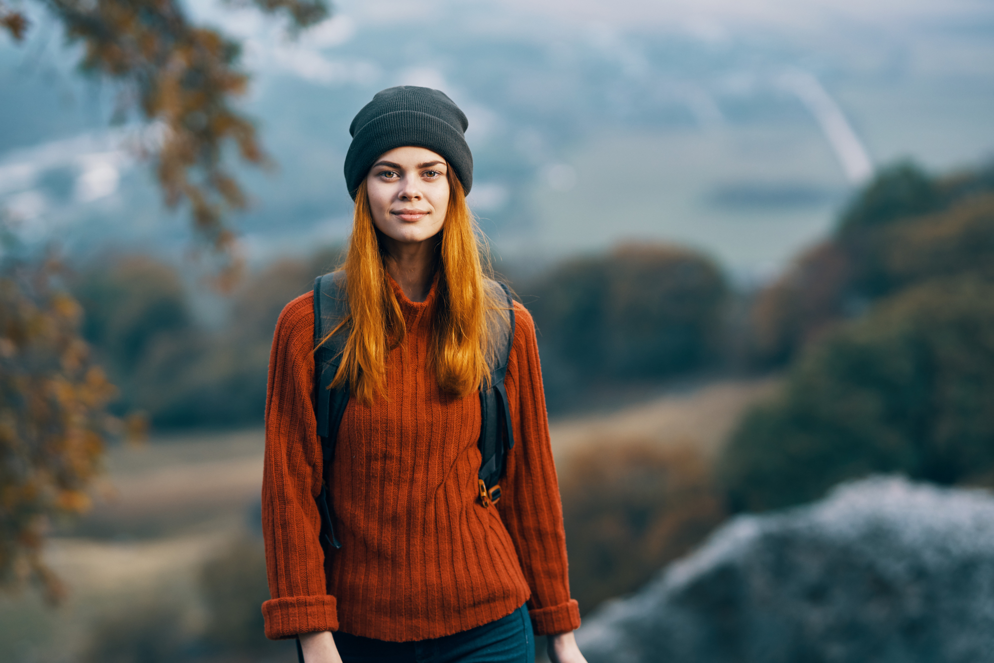 A young person with long red hair wearing a green beanie and rust-colored sweater stands outdoors with a backpack on. The background features a blurred natural landscape with trees and mountains.