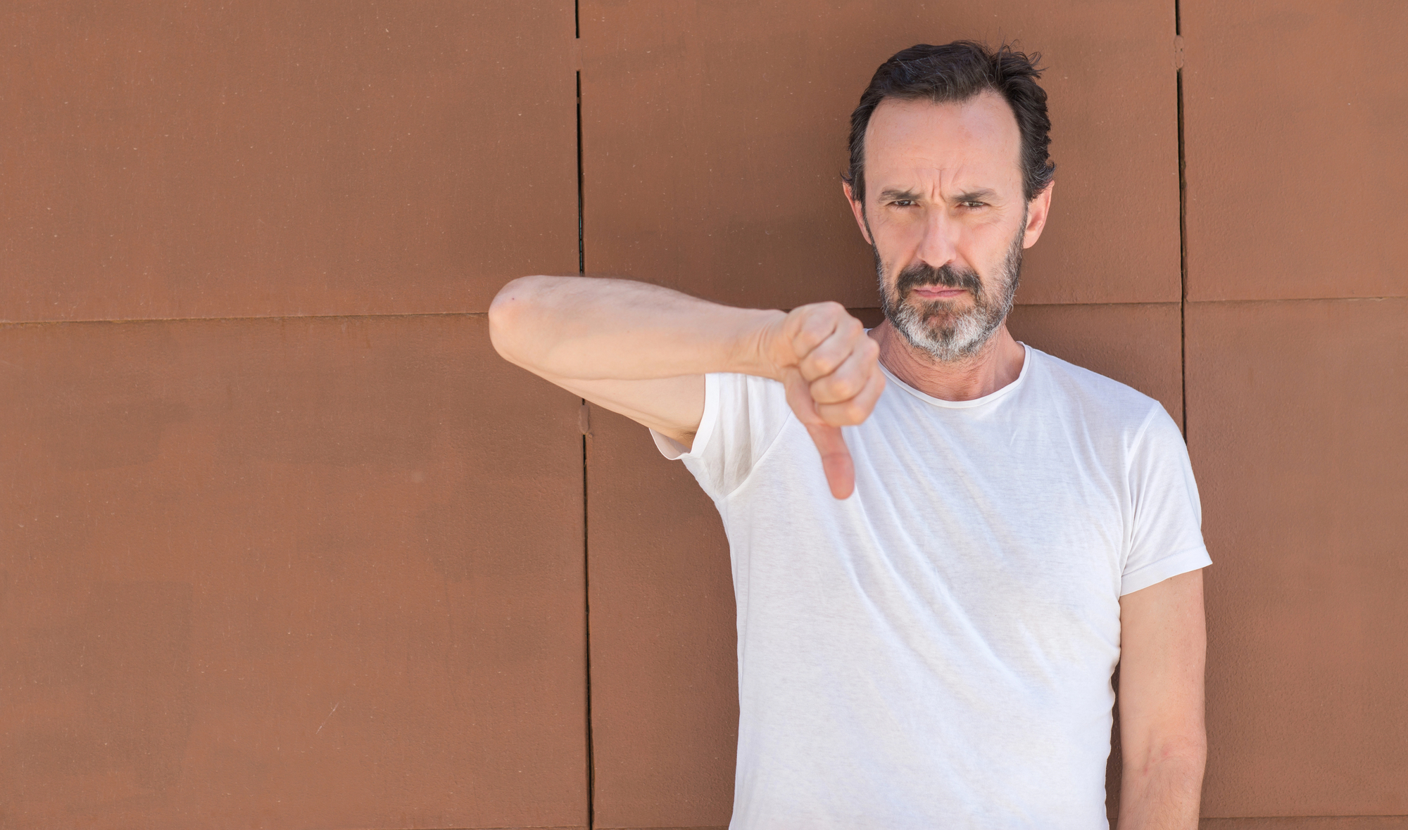 A man with short dark hair and a beard stands against a brown wall, wearing a plain white t-shirt. His expression is serious, and he is giving a thumbs-down gesture with his right hand.