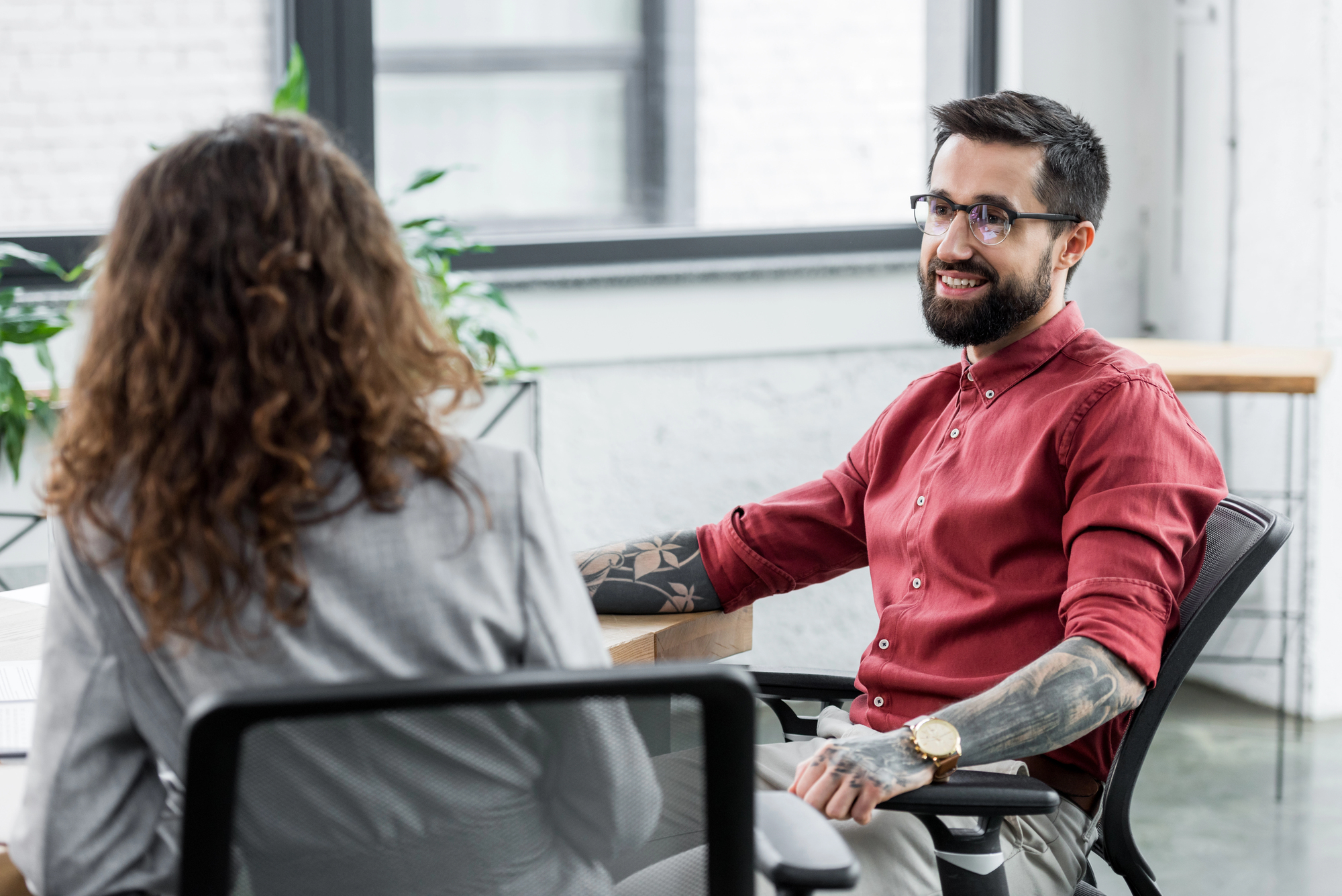 A man with glasses, a beard, and tattoos on his arm, wearing a red shirt, is smiling while sitting in a modern office. He is facing a woman with curly hair in a gray suit, who is turned away from the camera. They are seated at a wooden desk.