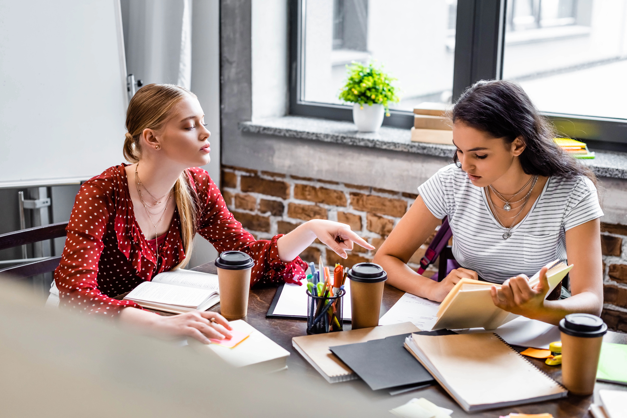 Two women sit at a table covered with notebooks, coffee cups, pens, and a potted plant. One woman, in a red polka dot blouse, points at the other woman's notebook. The other woman, in a striped shirt, looks down, appearing to study or read.