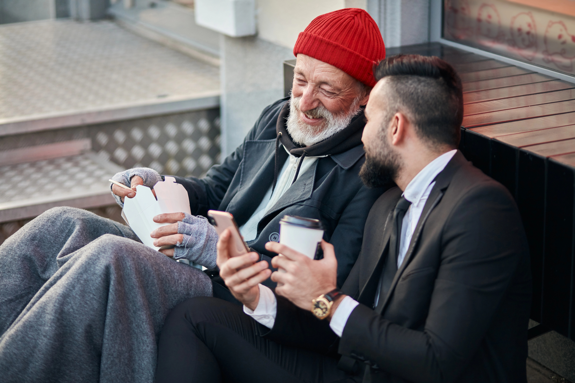 An older man wearing a red beanie and gloves, wrapped in a blanket, sits on the street smiling and talking with a younger man in a suit holding a coffee cup. The younger man shows something on his phone, and they appear to be enjoying a friendly conversation.