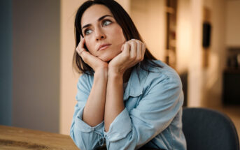 A woman with long dark hair and wearing a light denim shirt rests her chin on her hands and gazes thoughtfully into the distance while sitting at a wooden table in a softly lit room.