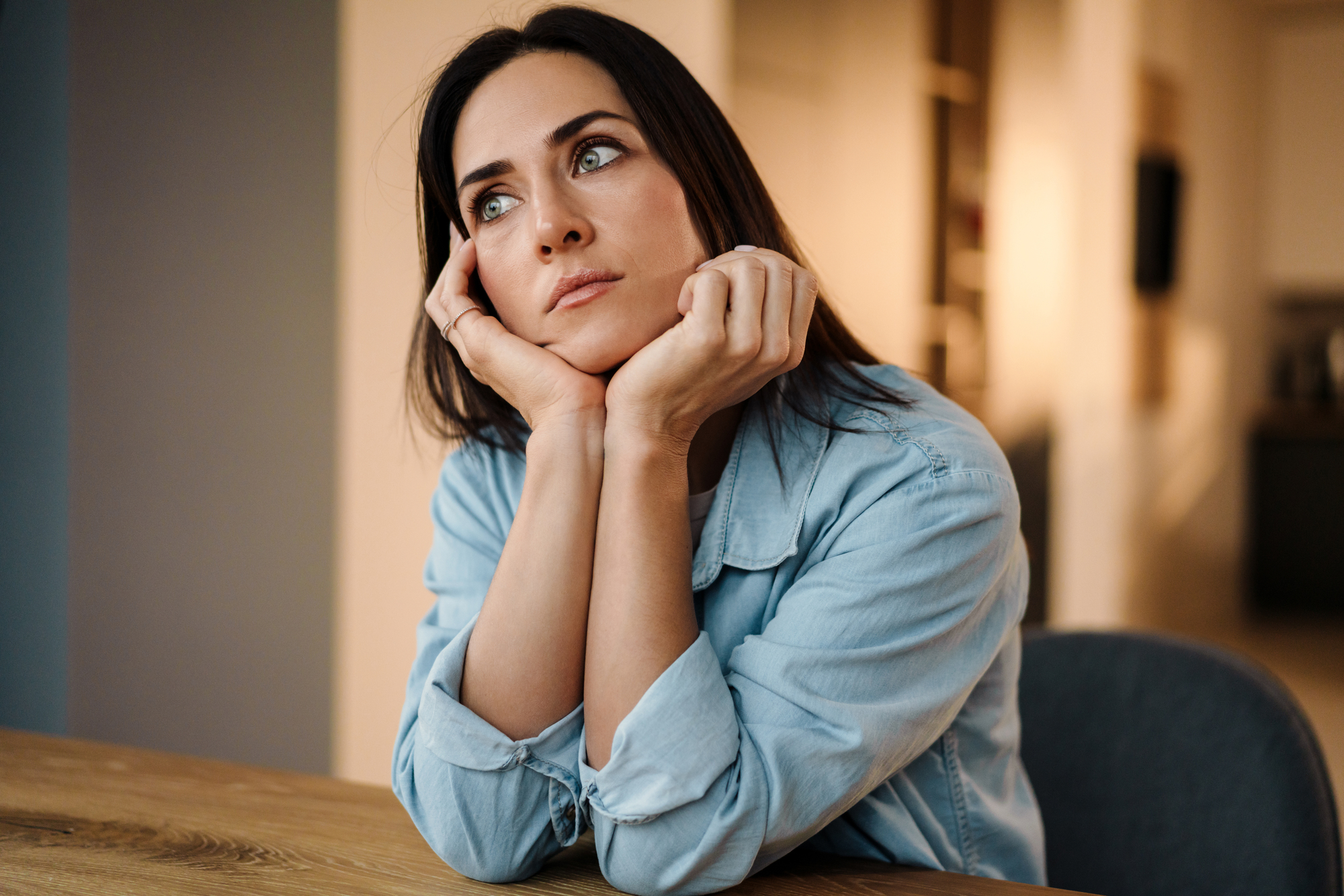 A woman with long dark hair and wearing a light denim shirt rests her chin on her hands and gazes thoughtfully into the distance while sitting at a wooden table in a softly lit room.