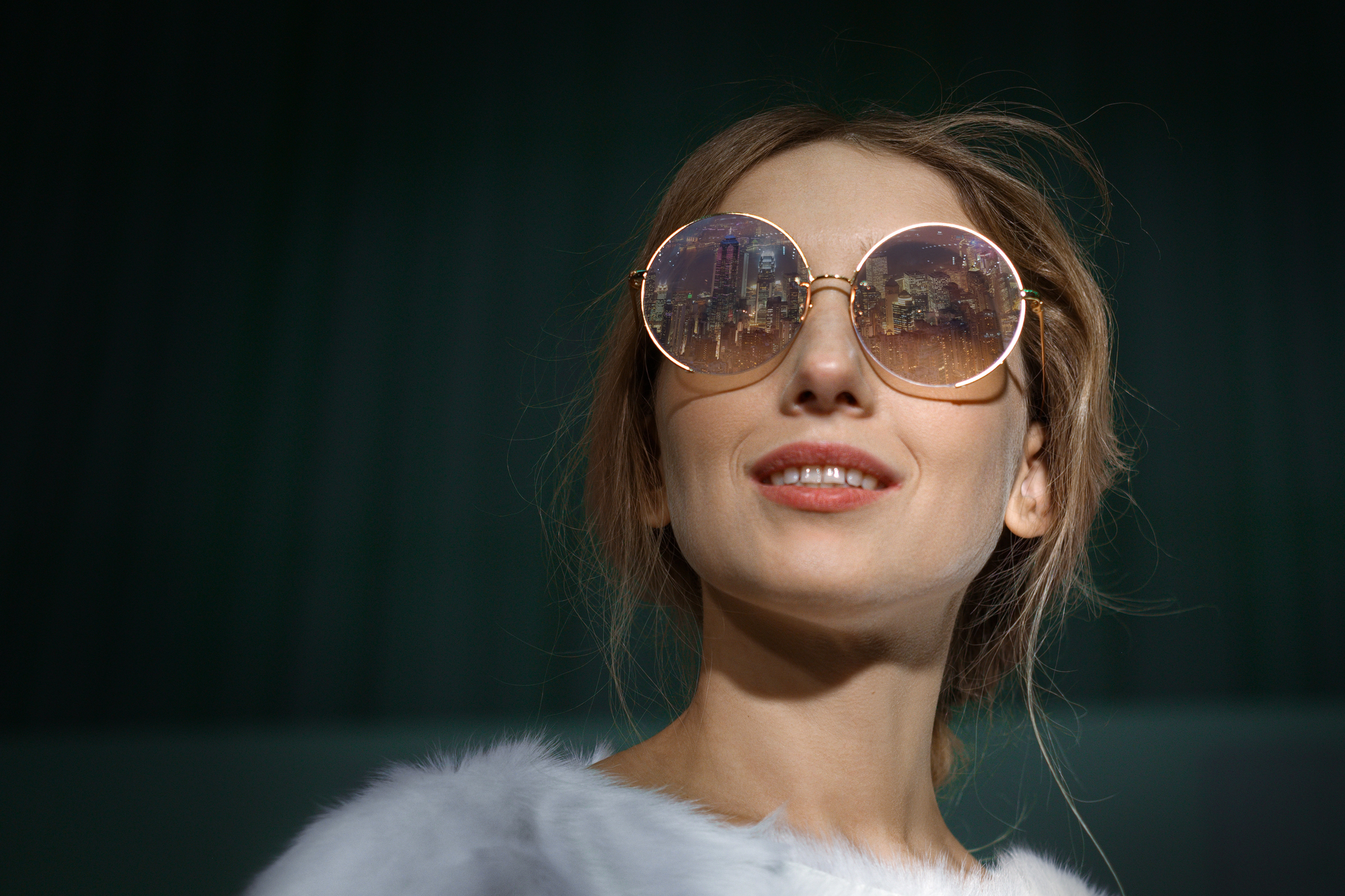 A woman with light brown hair, wearing large round sunglasses that reflect a city skyline at night. She has a slight smile and is dressed in a fluffy white top. The background is dark green, giving a contrast to her lighter attire and accessories.