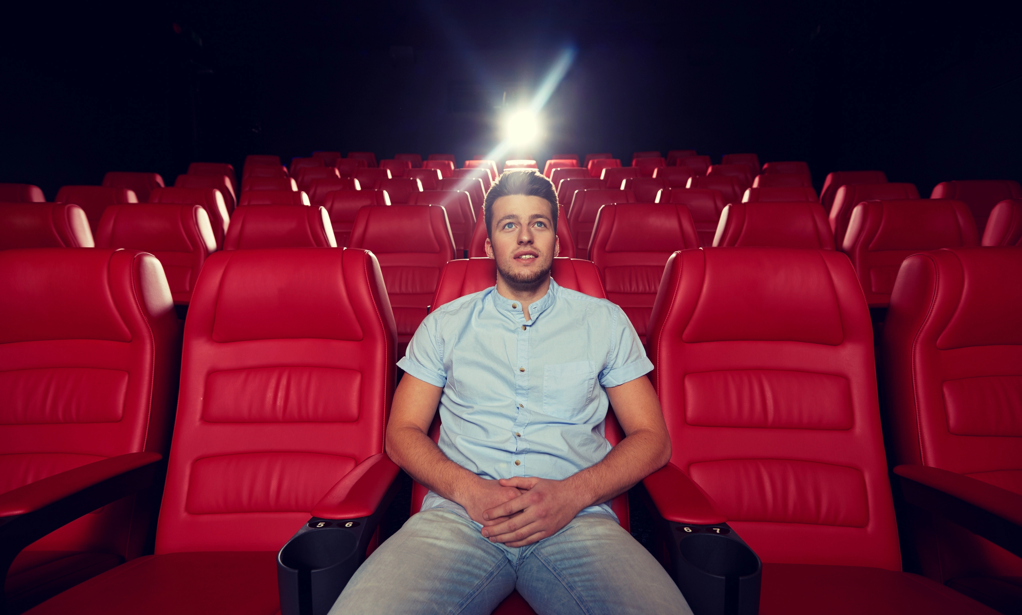 A young man wearing a light blue shirt and jeans sits alone in the middle of a theater, surrounded by rows of empty red seats. A bright projector light shines from the back, casting a glow on him.