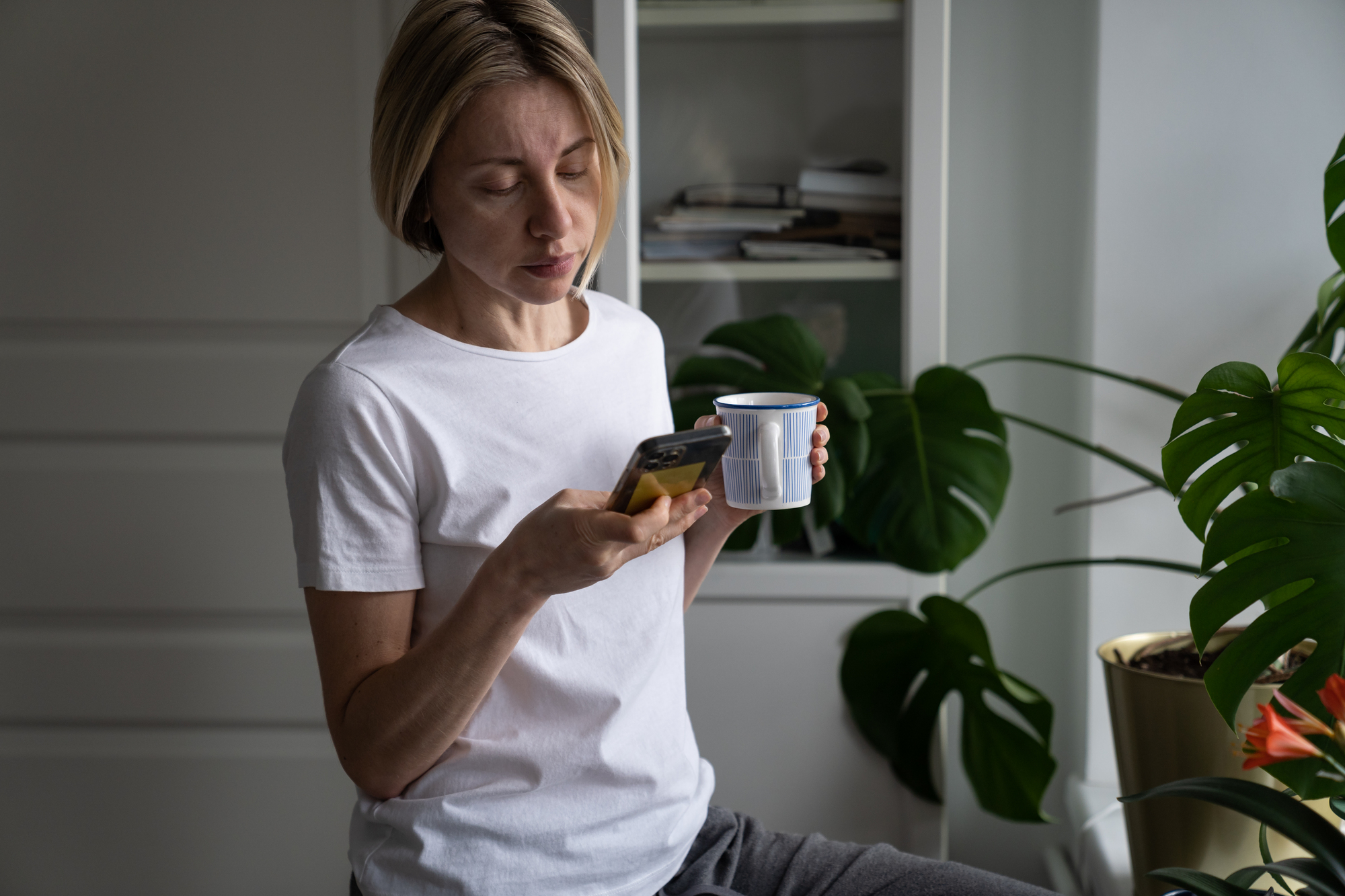 A person with short blonde hair is sitting and looking at a smartphone while holding a white cup. They are wearing a plain white t-shirt. In the background, there are green houseplants and a white cabinet with books and folders.