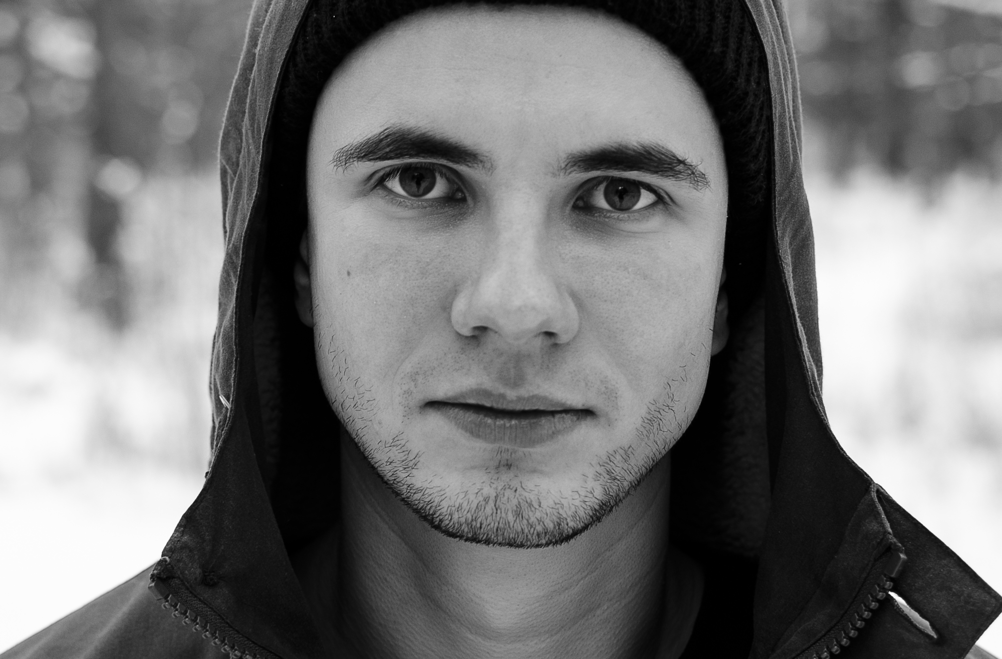 Black and white close-up of a young man looking directly at the camera. He has a slight smile, short beard, and is wearing a hooded jacket and beanie. The background is blurred.