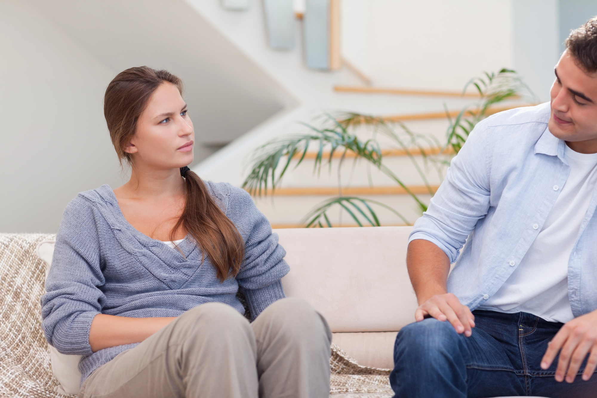 A woman with long brown hair, wearing a grey sweater, sits on a beige couch, looking at a man beside her. The man, with dark hair, is dressed in a light blue shirt and jeans, and appears to be speaking to her. They are in a modern living room.