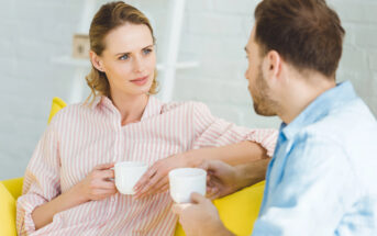 A woman and a man sitting on a yellow couch, holding white mugs and engaged in conversation. The woman has blonde hair, and is wearing a pink striped shirt, while the man has brown hair and is wearing a light blue shirt. They are indoors with a white background.