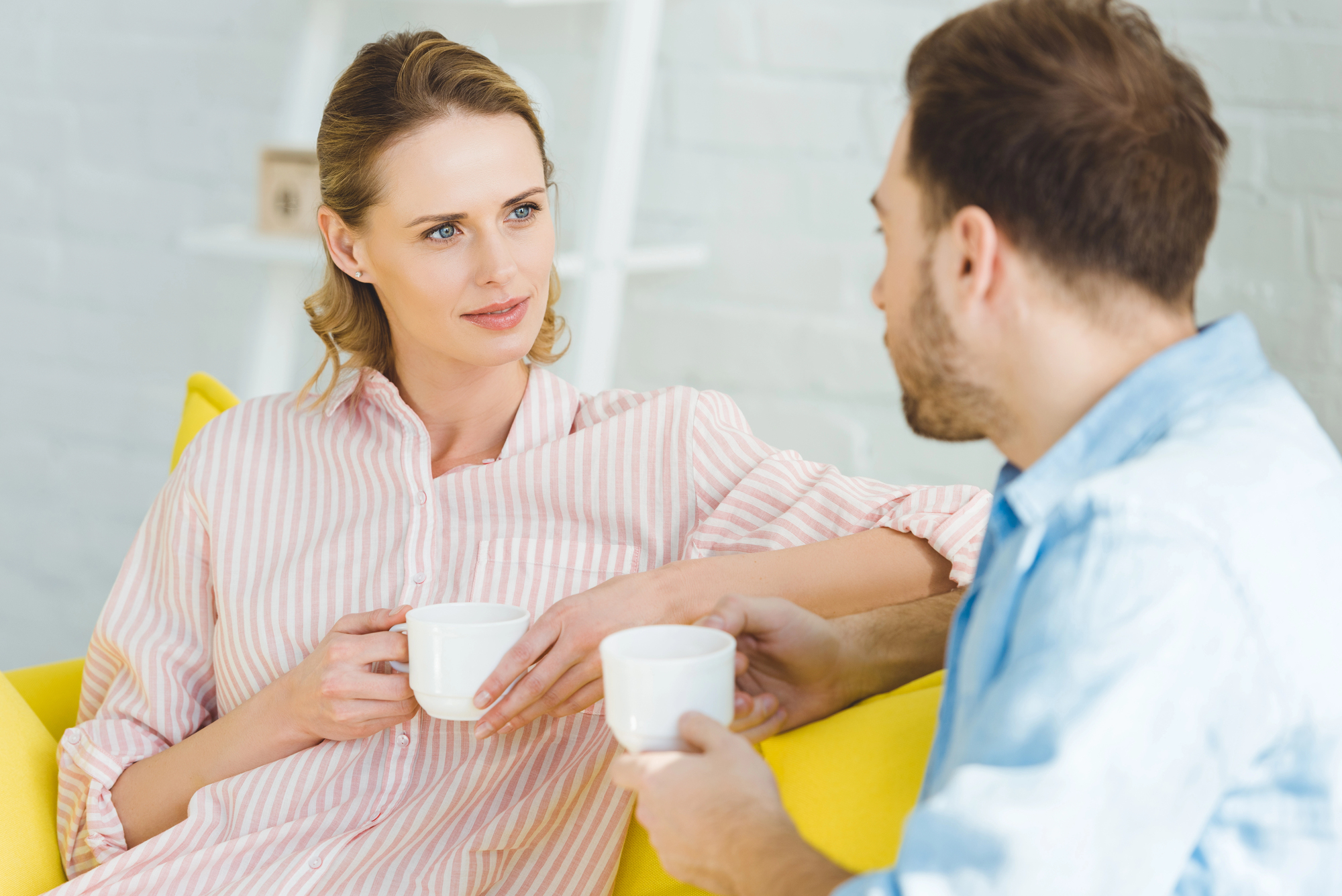 A woman and a man sitting on a yellow couch, holding white mugs and engaged in conversation. The woman has blonde hair, and is wearing a pink striped shirt, while the man has brown hair and is wearing a light blue shirt. They are indoors with a white background.