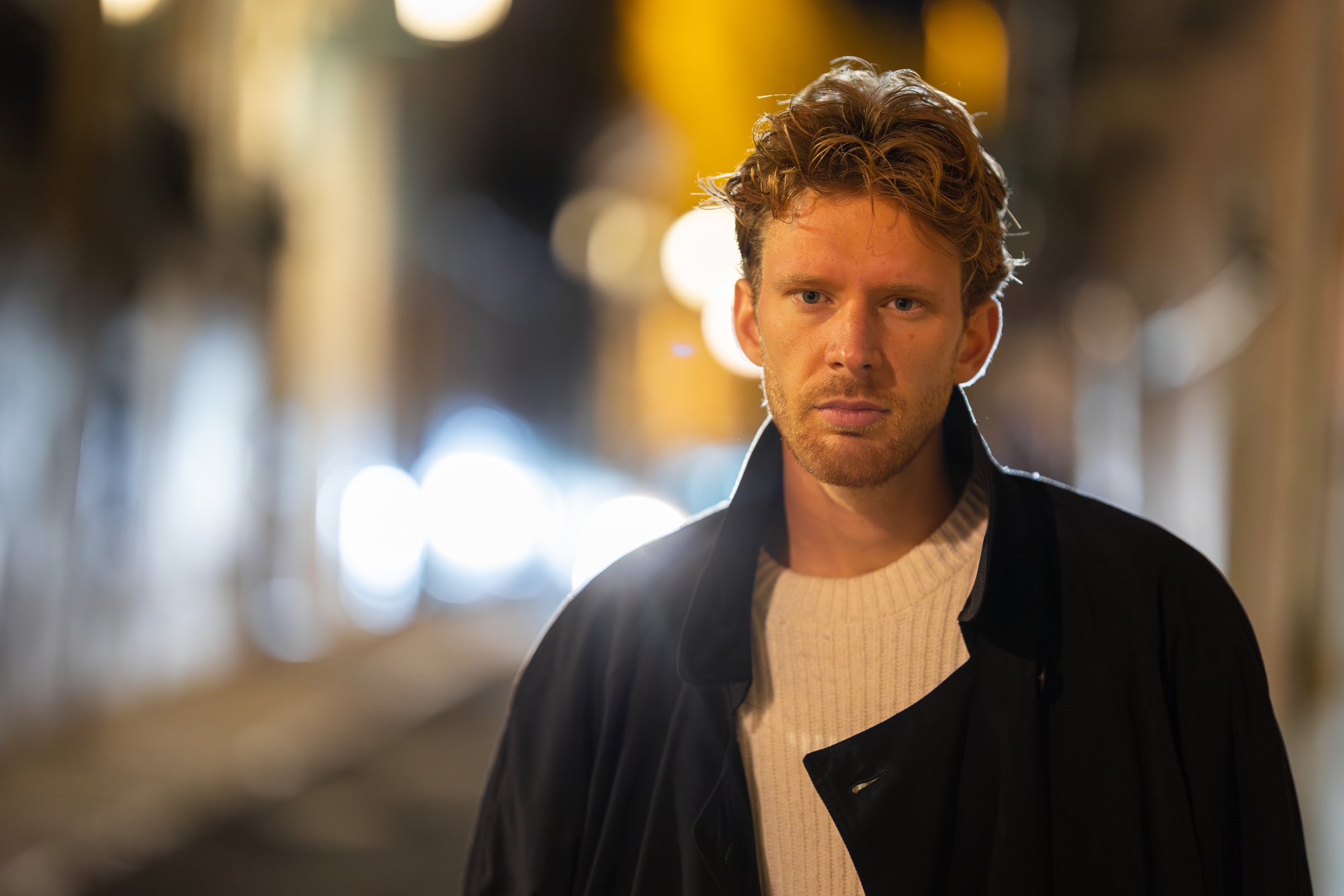 A man with wavy hair stands in the middle of a dimly lit street at night. He wears a white sweater and a dark coat. The background is blurry with illuminated streetlights and abstract light effects. His expression is neutral.