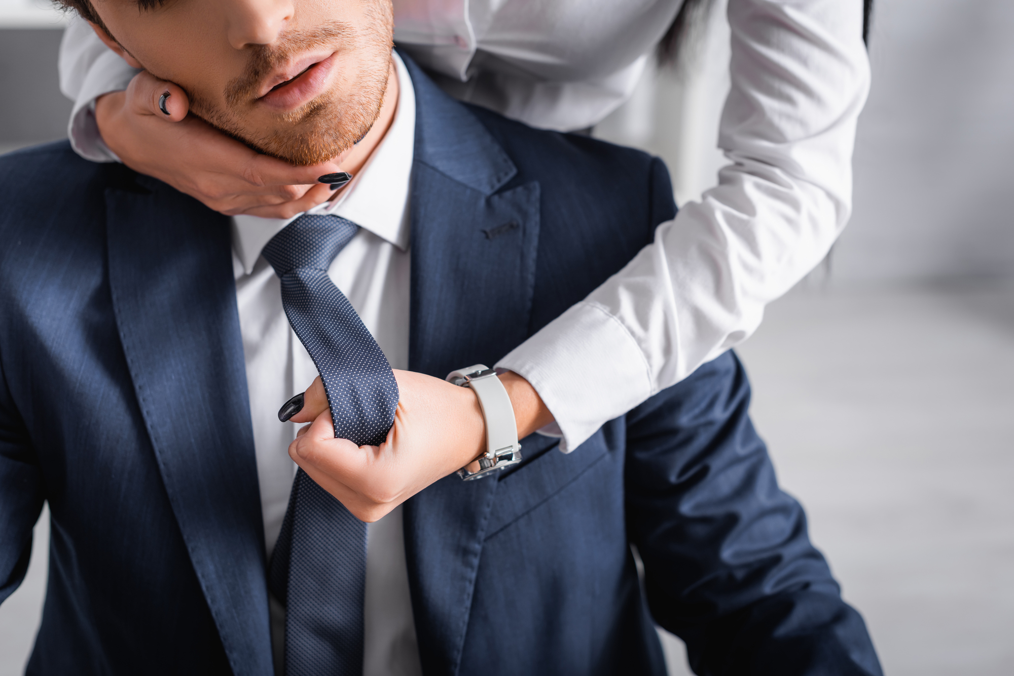 A person in a blue suit and tie is seated while another person, wearing white long sleeves, stands behind and grabs the seated person's tie with one hand. The background is blurred.