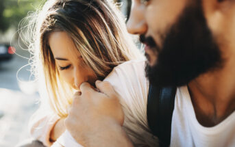 A woman with long, blonde hair gently kisses the shoulder of a bearded man who is holding her close. They both appear to be outdoors, sharing an intimate and affectionate moment. The background is slightly blurred, emphasizing their connection.